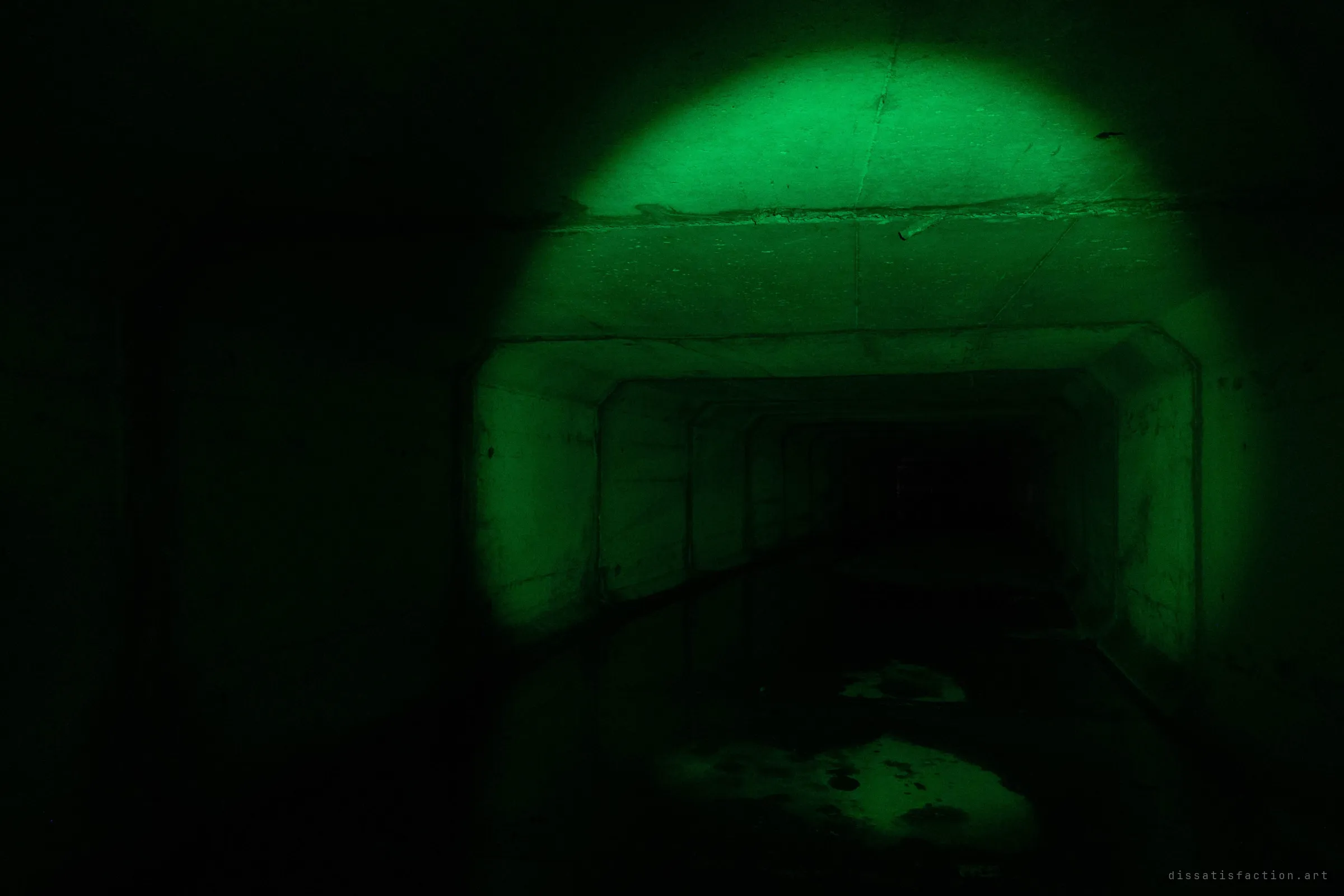A single green beam picks out the mouth of a square concrete drain, its stepped segments falling away into black. Most of the frame stays dark, with only wet patches on the floor catching the light.