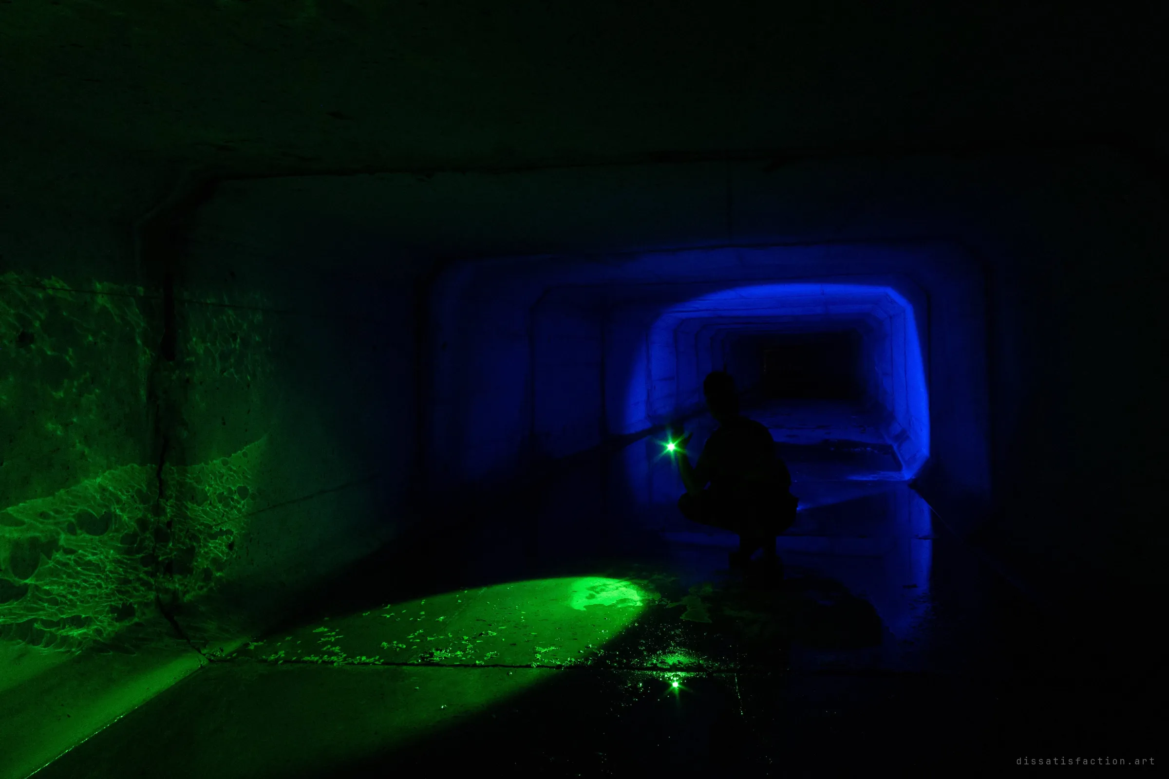A crouched silhouette holds a small green light in a square storm drain, with a blue beam lighting the receding concrete segments behind them. Green light ripples across the left wall and pools on the wet floor.