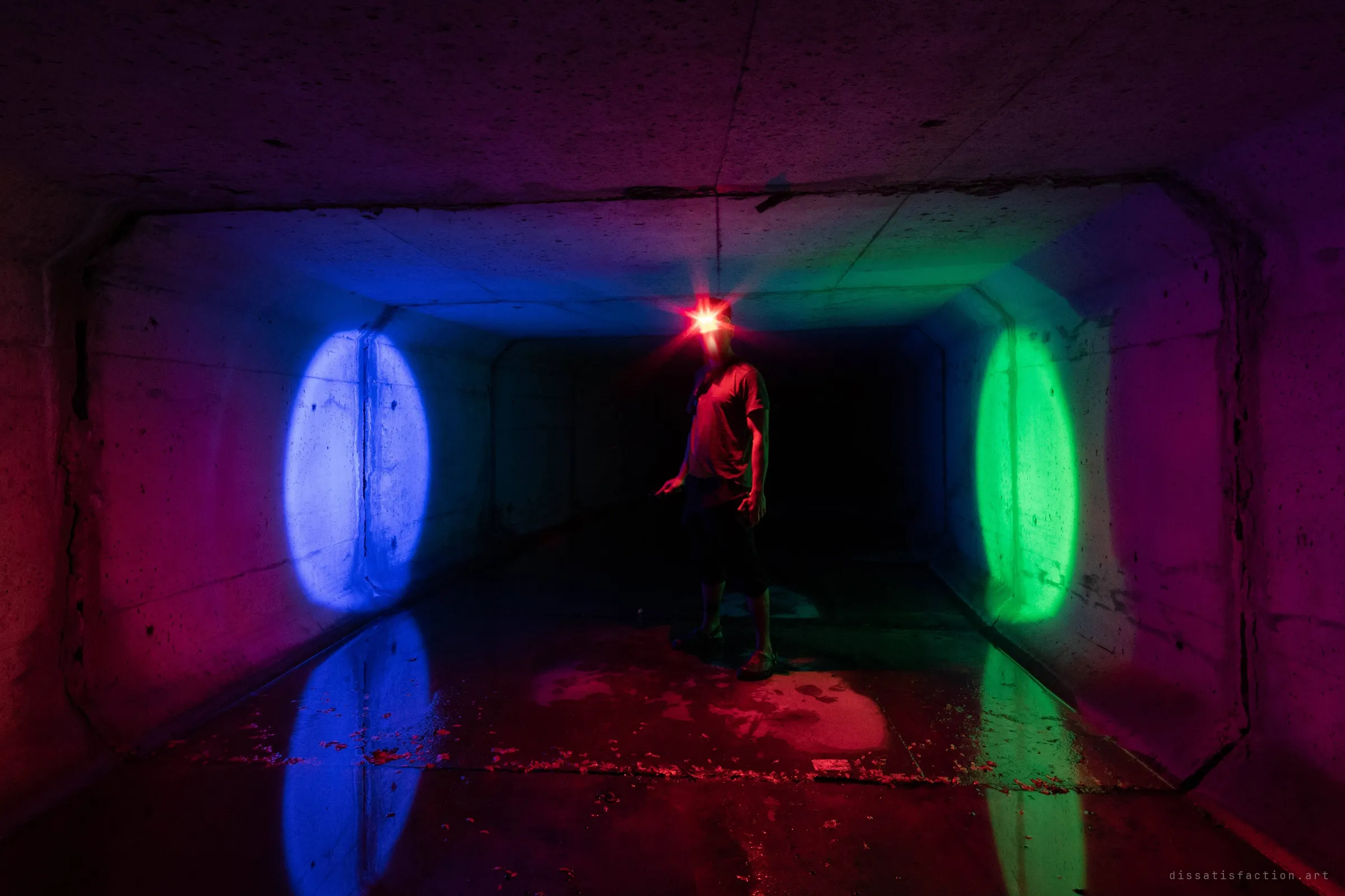 A figure stands in the middle of a dark concrete box culvert, a red headlamp blazing where their face would be. Blue and green spotlights cast two oval pools on the side walls, both mirrored in the wet floor, turning the plain drain into a colour-split stage.