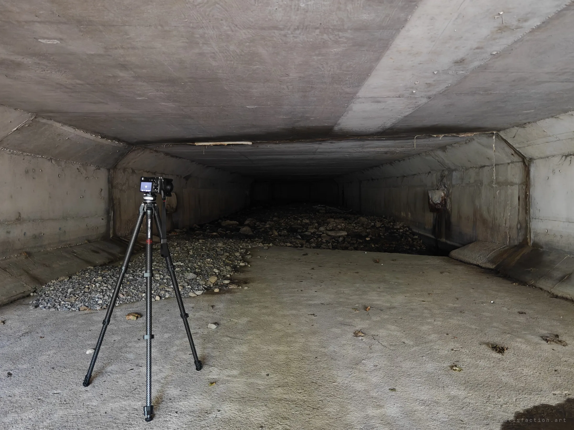 A camera on a carbon tripod at the mouth of a wide, flat-roofed concrete box culvert, with a bed of river stones running back into the dark. It shows how wide and low the drain is.