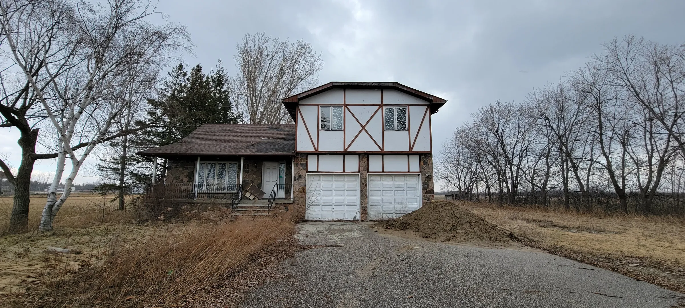 A shuttered split-level house with mock-Tudor timbering and a fieldstone base stands at the end of a cracked driveway, a dirt pile blocking one garage door. Bare birches and dead grass set the late-winter mood of the abandoned family home.