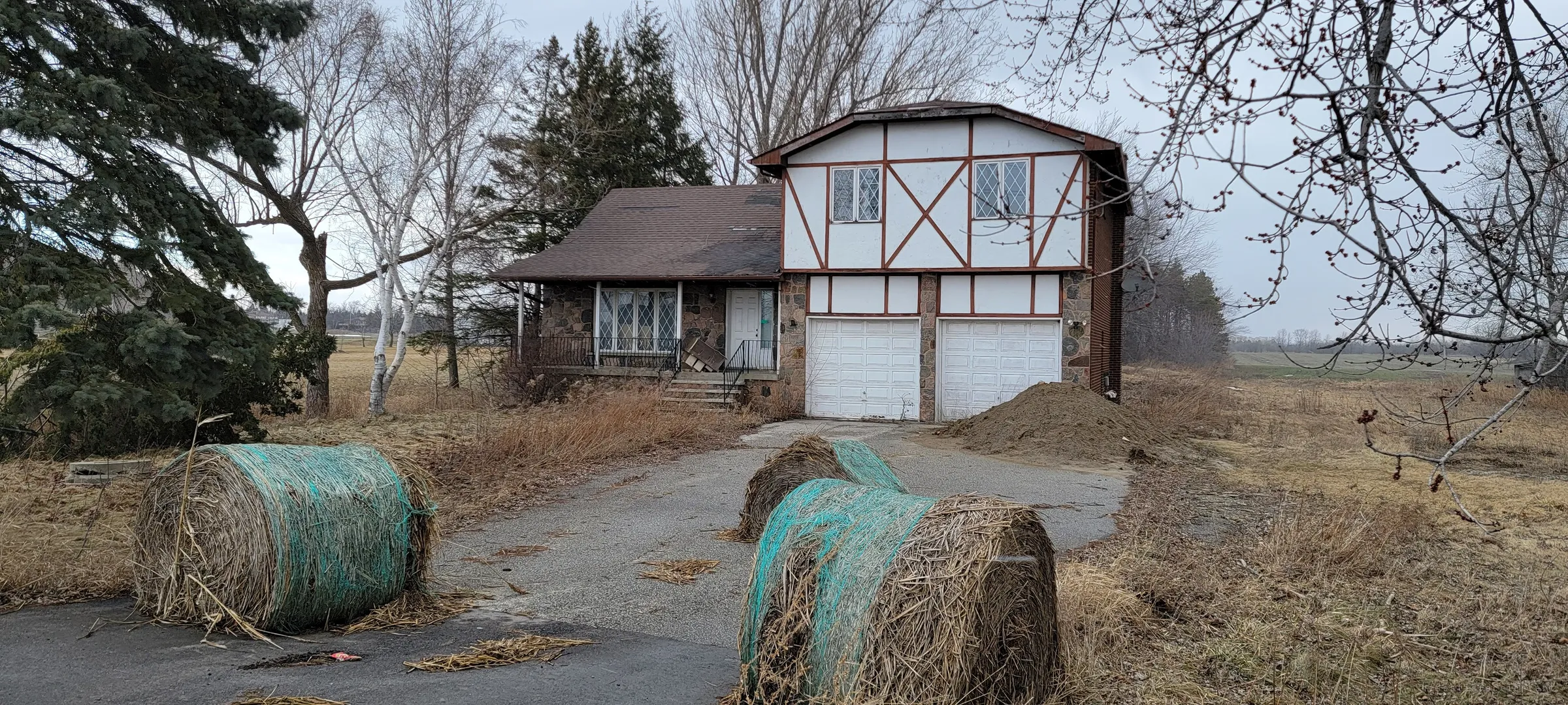 Round hay bales wrapped in green netting sit across the driveway in front of the mock-Tudor house, blocking the way in. The barricade shows how the property was sealed off after it was left.
