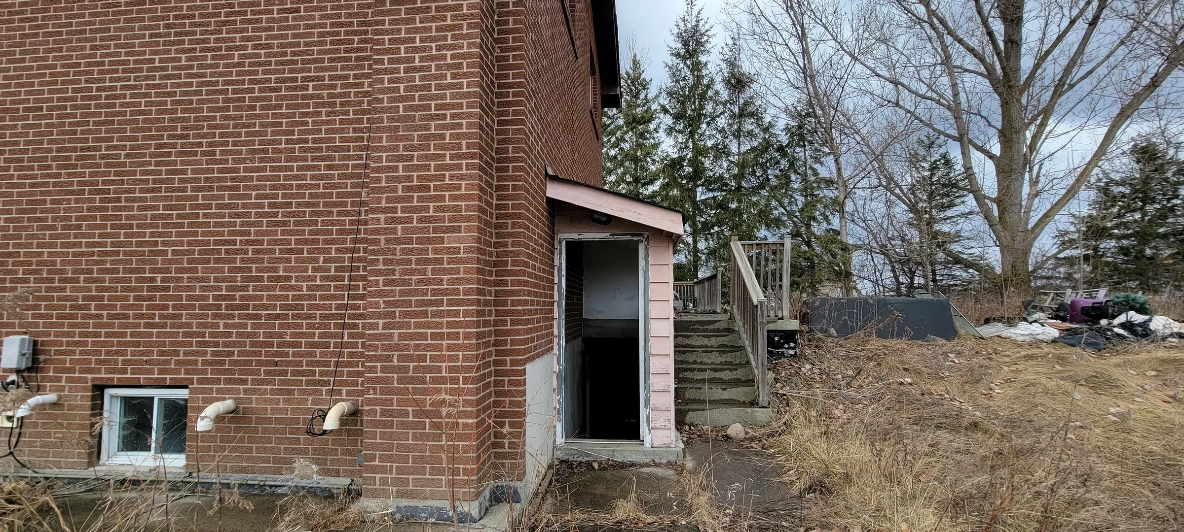 A small pink-sided porch on the brick side wall opens onto a dark doorway down to the basement, next to a wooden stair and a yard of dumped junk. It's one of the ways into the empty house.