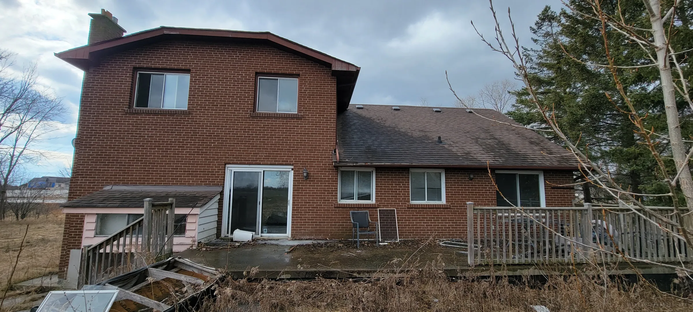 The brick back of the house with a sliding patio door, a railed wooden deck and a cracked concrete patio littered with a chair and a fallen screen. Seen from the overgrown yard, the family's outdoor space has been left to rot.