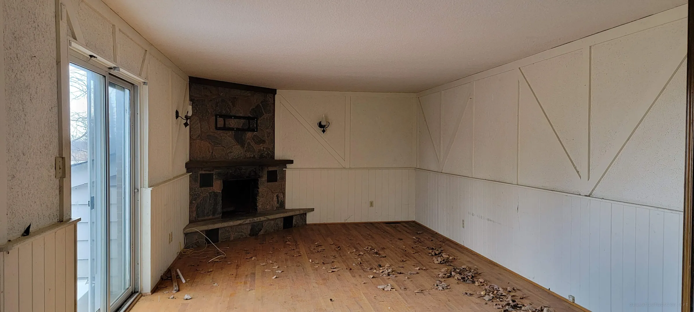 An empty living room with a corner fieldstone fireplace, iron wall sconces and cross-braced panelling, dead leaves scattered on the hardwood floor. The dated decor still shows how the family room once looked.