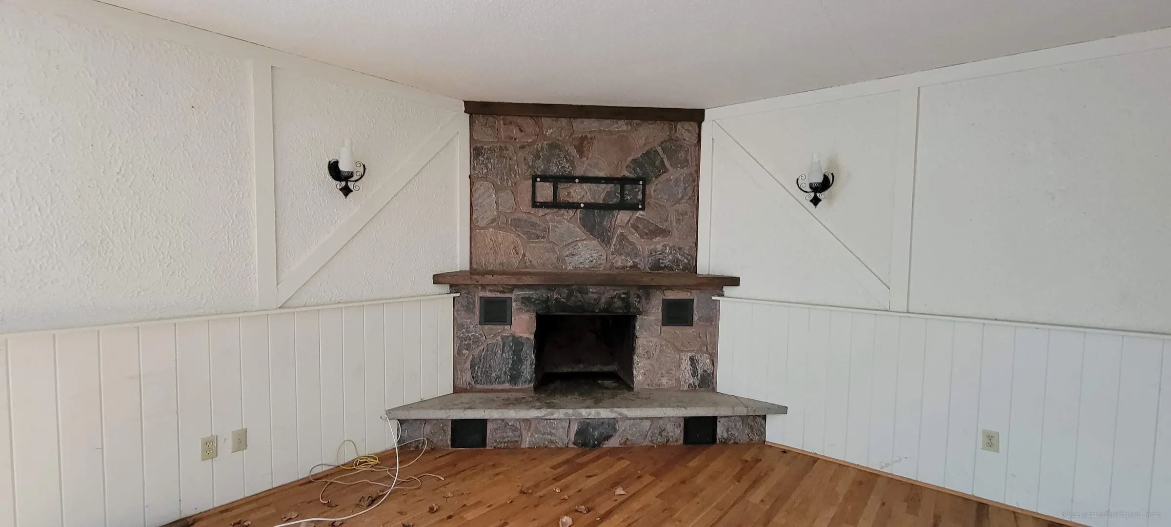 A close, square-on view of the corner fieldstone fireplace, its empty TV bracket above the mantel and matching iron sconces on either side. A loose cable and a few leaves lie on the floor where furniture used to be.