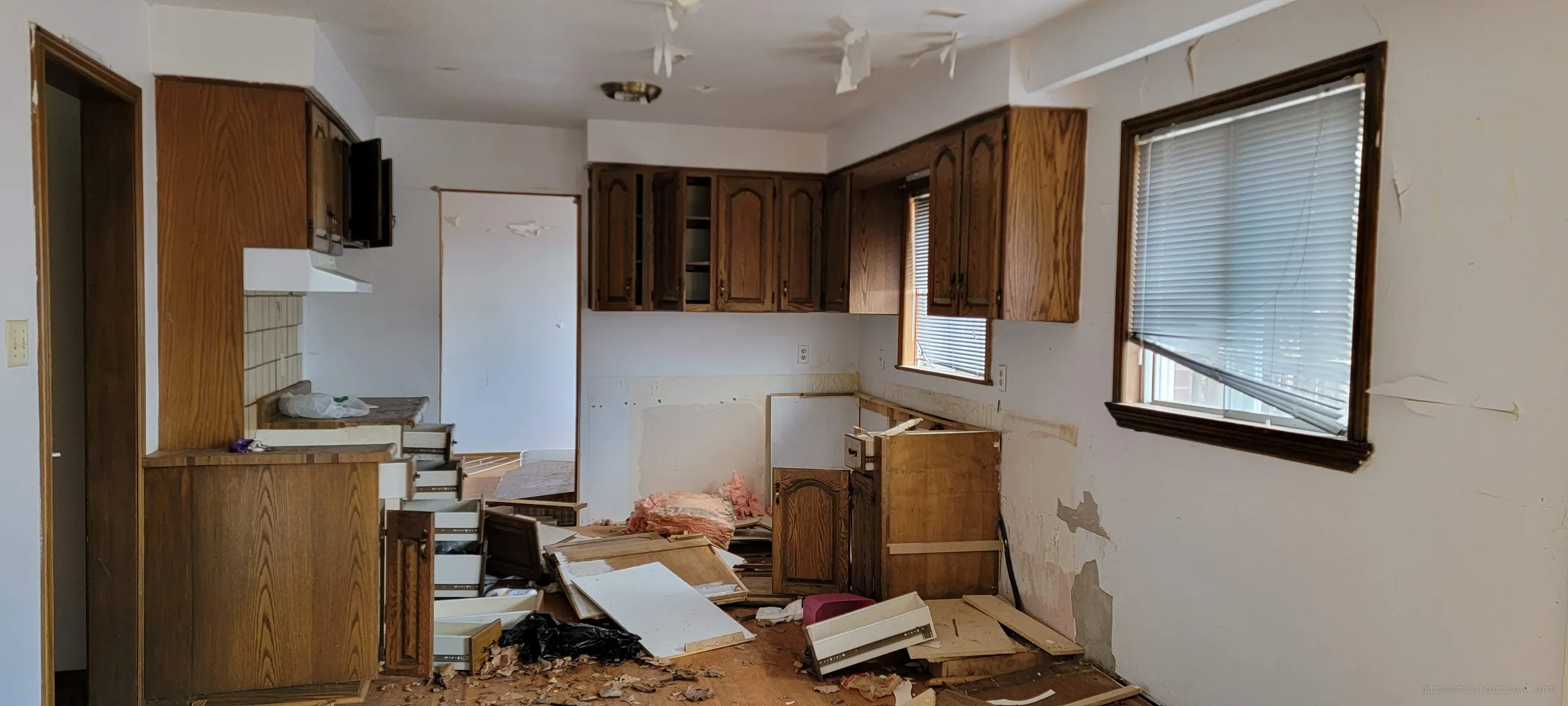 A gutted kitchen with oak cabinets still on the walls, the lower units torn out and their drawers and doors piled on the floor with pink insulation. Ceiling paper hangs in strips above the wreckage.