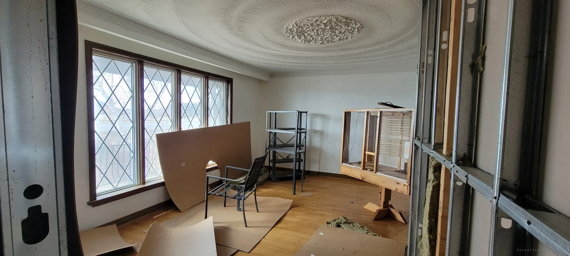 A front room under a swirled plaster ceiling medallion, with leaded diamond windows, fibreboard sheets on the floor, a metal shelf and a large wooden bird cage on a stand. Exposed steel studs frame the doorway, halfway through a renovation that never finished.