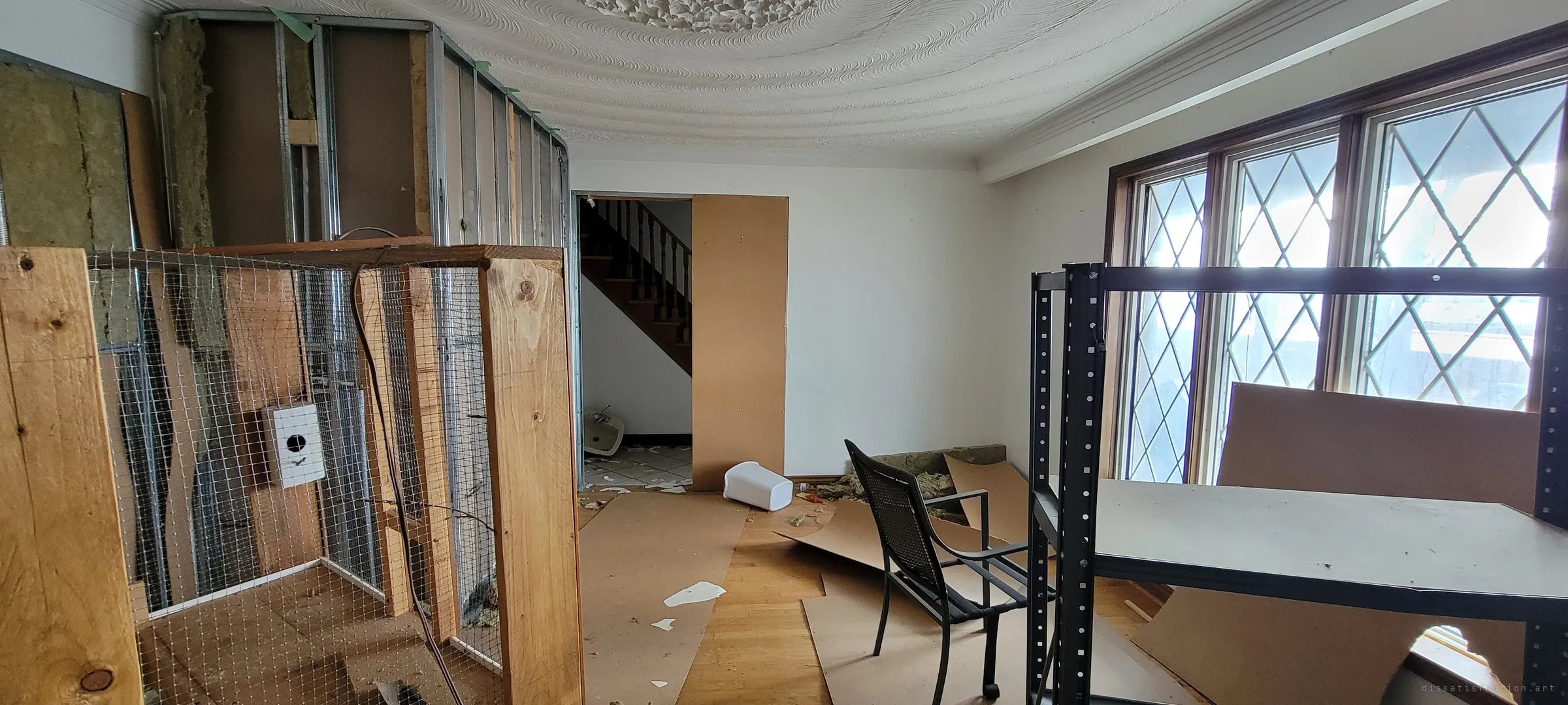 The same swirl-ceilinged room seen from the other side: a mesh-and-timber enclosure against a half-stripped stud wall, a patio chair and shelving by the bright leaded windows, and a doorway to the stairs beyond. The unfinished renovation left the room part home, part building site.