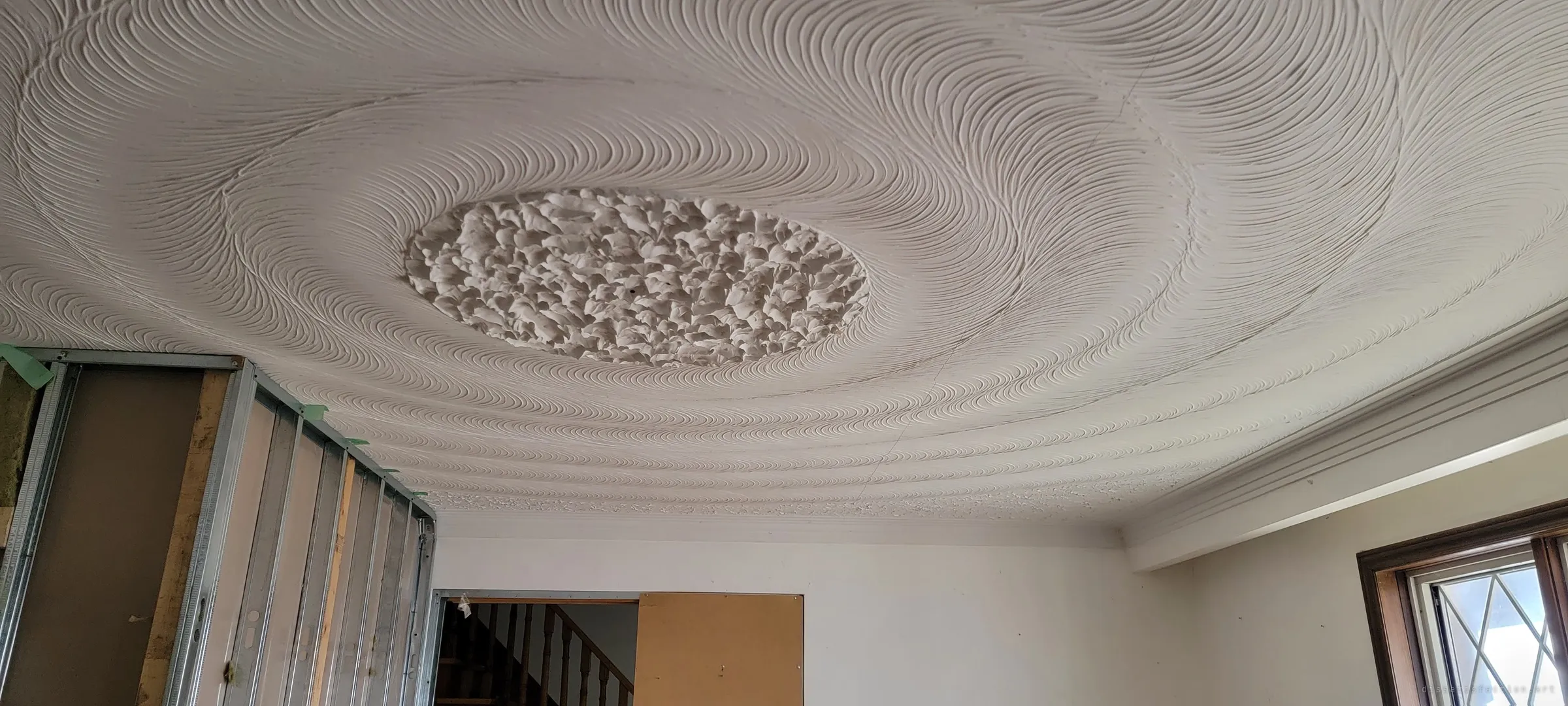 A sweeping hand-combed plaster ceiling spirals around a rough stippled centre, above a room half stripped back to bare steel studs. The ornate finish sits against the renovation that stalled mid-way.