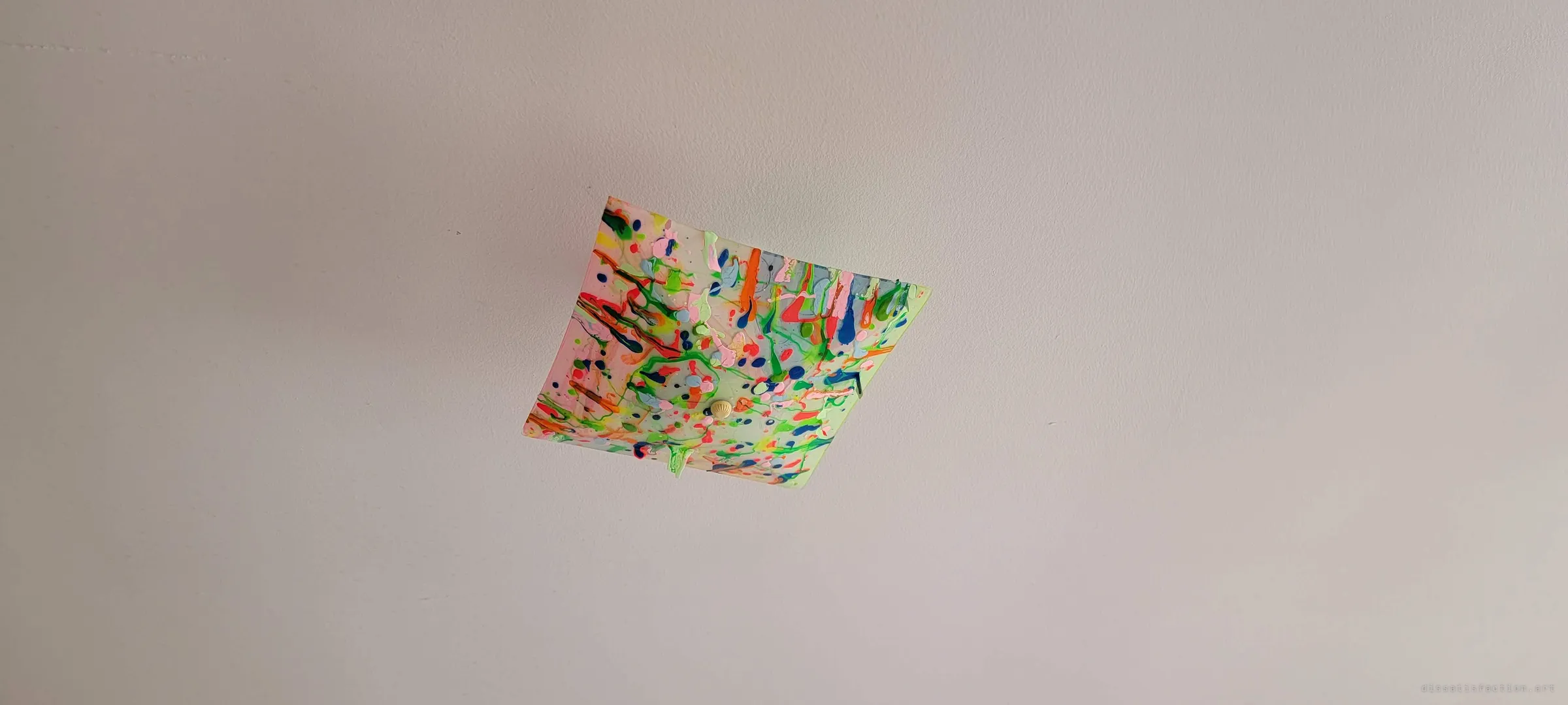 A square ceiling light shade splattered with neon paint drips hangs on a plain white ceiling. A child's playful touch left behind in an empty room.