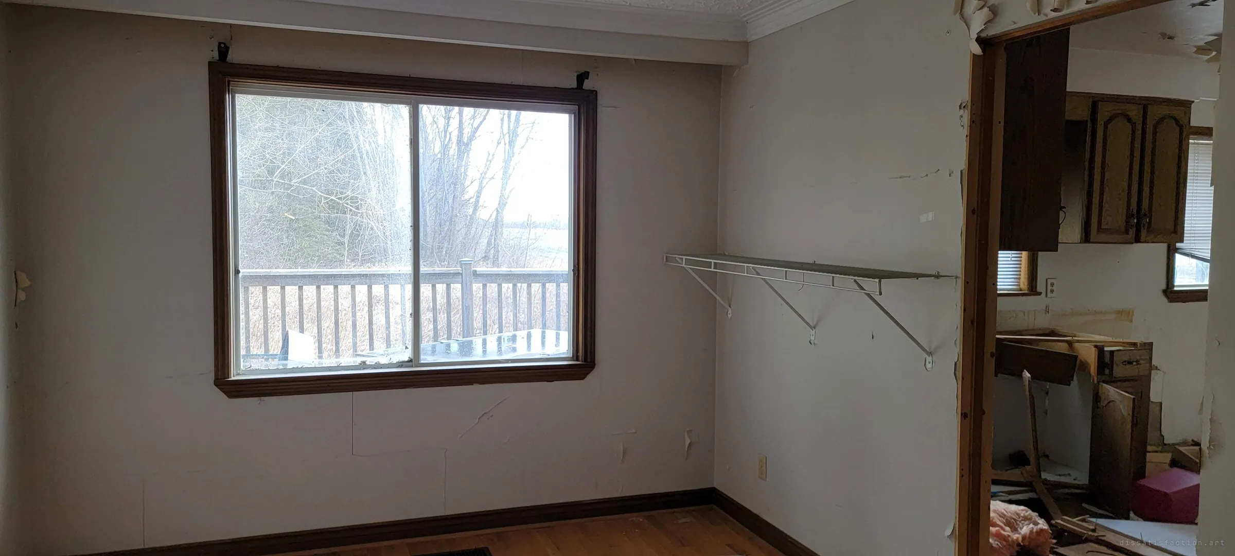 A bare room with a wire shelf and peeling walls looks out through a large window onto a wooden deck, leafless trees and open fields. Through the doorway, a gutted kitchen with torn-out cabinets.