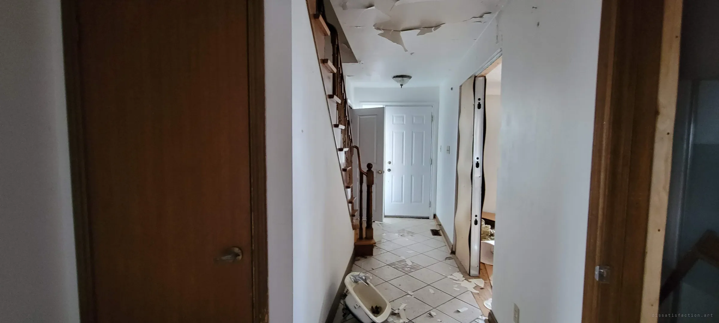 A narrow hallway runs past the staircase to the front door, its ceiling peeling in sheets and a broken sink lying on the debris-strewn tile floor. The ultra-wide view frames it between two doorways.