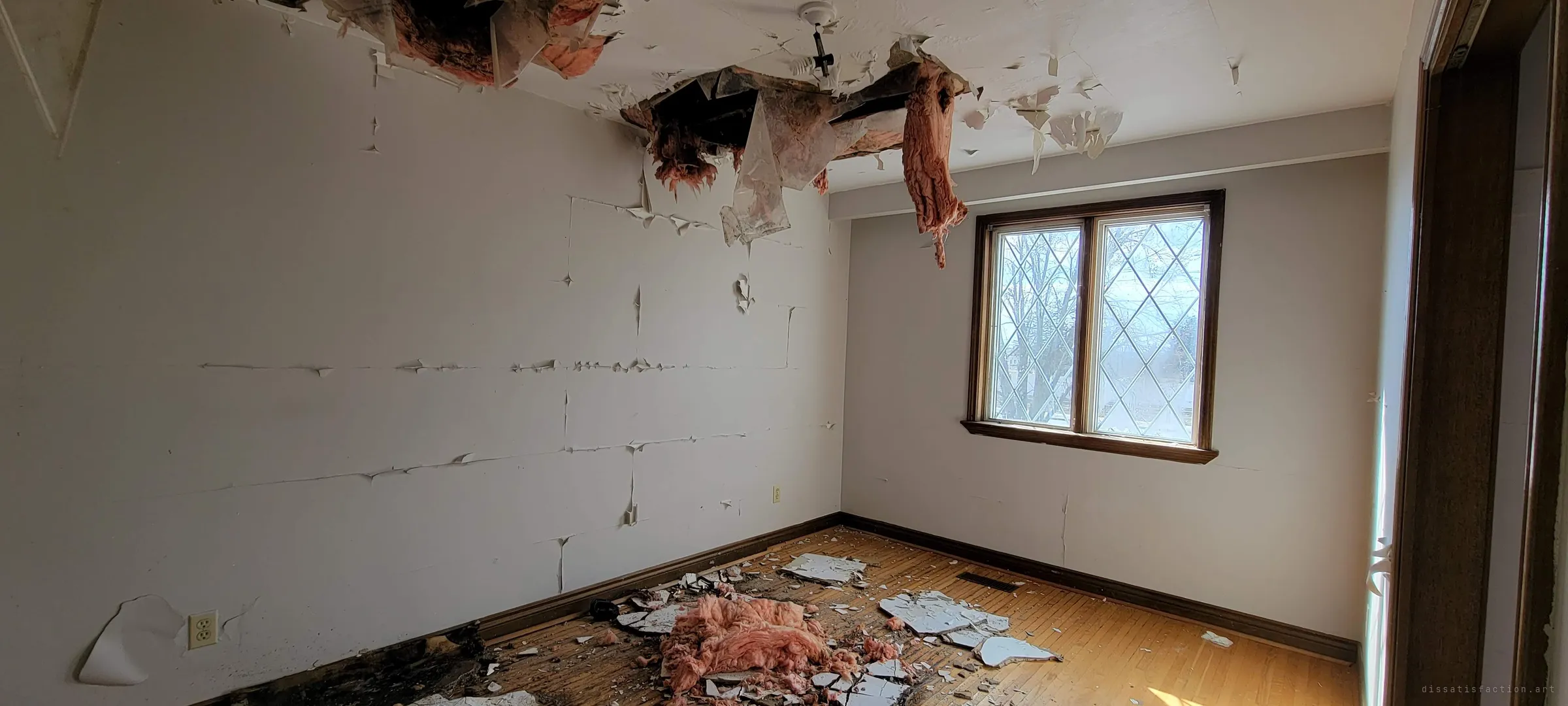 Pink insulation spills through a hole torn in a bedroom ceiling and lies heaped on the floor beneath, while sunlight falls through a diamond-leaded window. The room is visibly caving in.