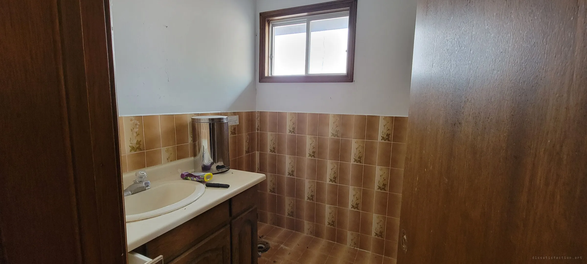 A small bathroom with floral brown tile, a cream sink and a steel bin, its toilet removed to leave a bare flange. The dated seventies finish is almost untouched.