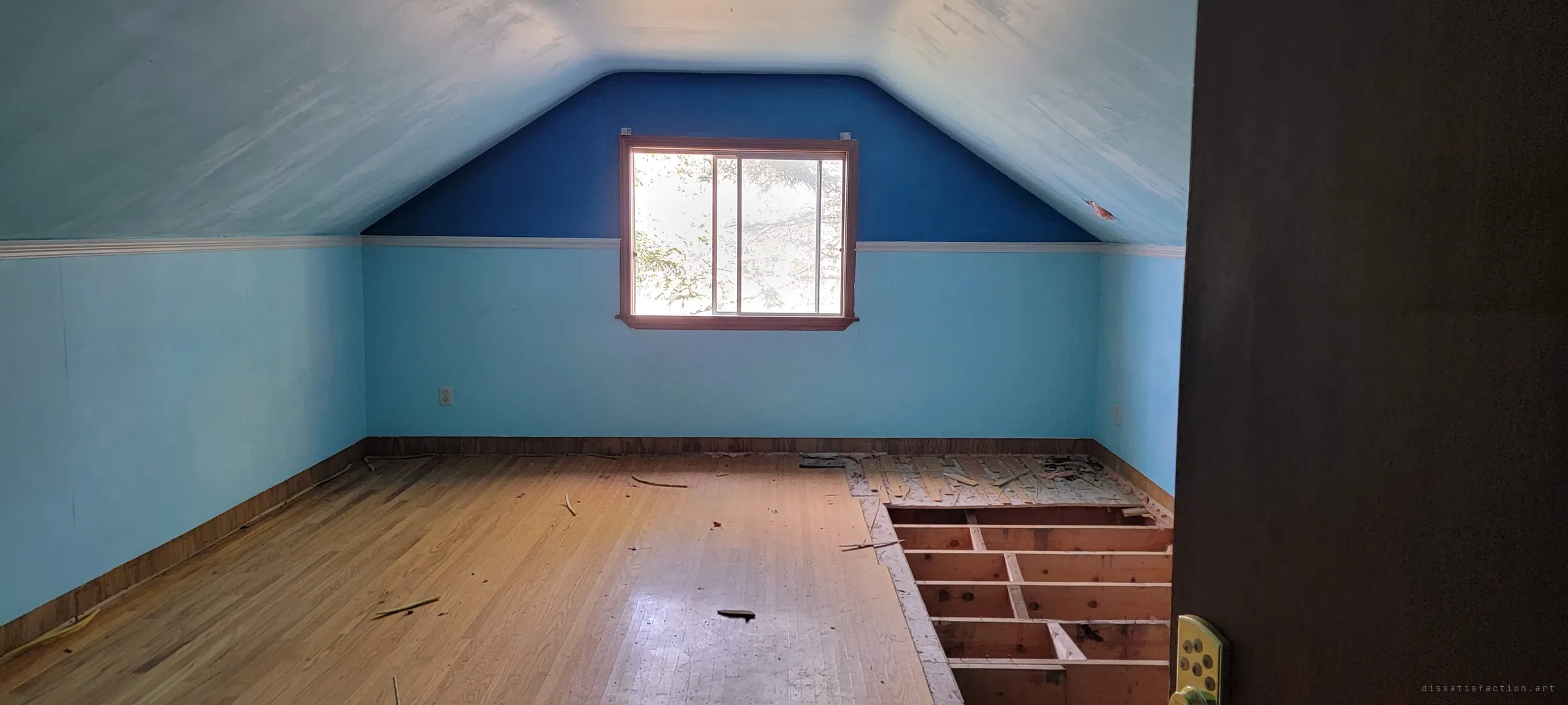 An attic bedroom painted in two shades of blue under a sloped ceiling, with part of the hardwood floor pulled up to expose the joists. A single window glows with trees outside.