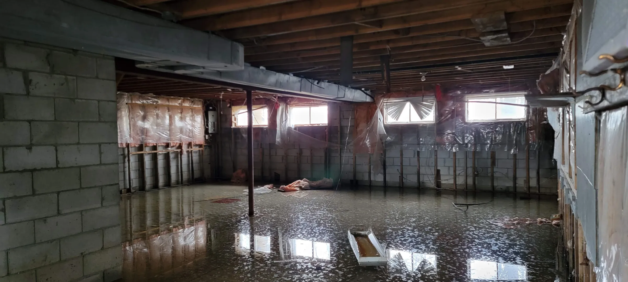 A stripped basement stands under a sheet of water, its insulation torn and plastic vapour barrier hanging loose. The small windows reflect in the flood across the floor.
