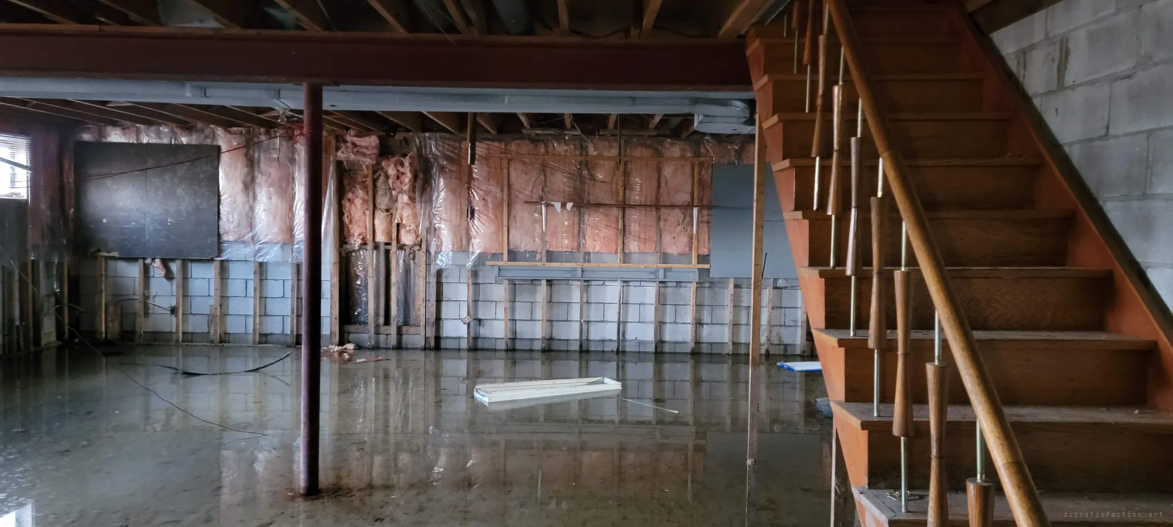 Another view of the flooded basement, from beside the wooden staircase with turned spindles. Pink insulation and bare studs line the walls behind the water.