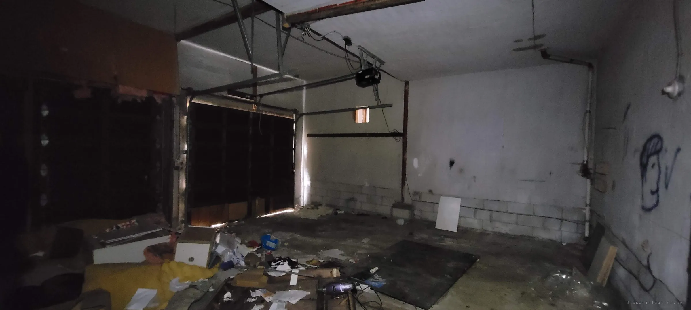 A dim garage with its door shut and the opener still hanging from the ceiling. The floor is covered in junk, loose papers and an old sofa, and a doodled face is sprayed on the wall.