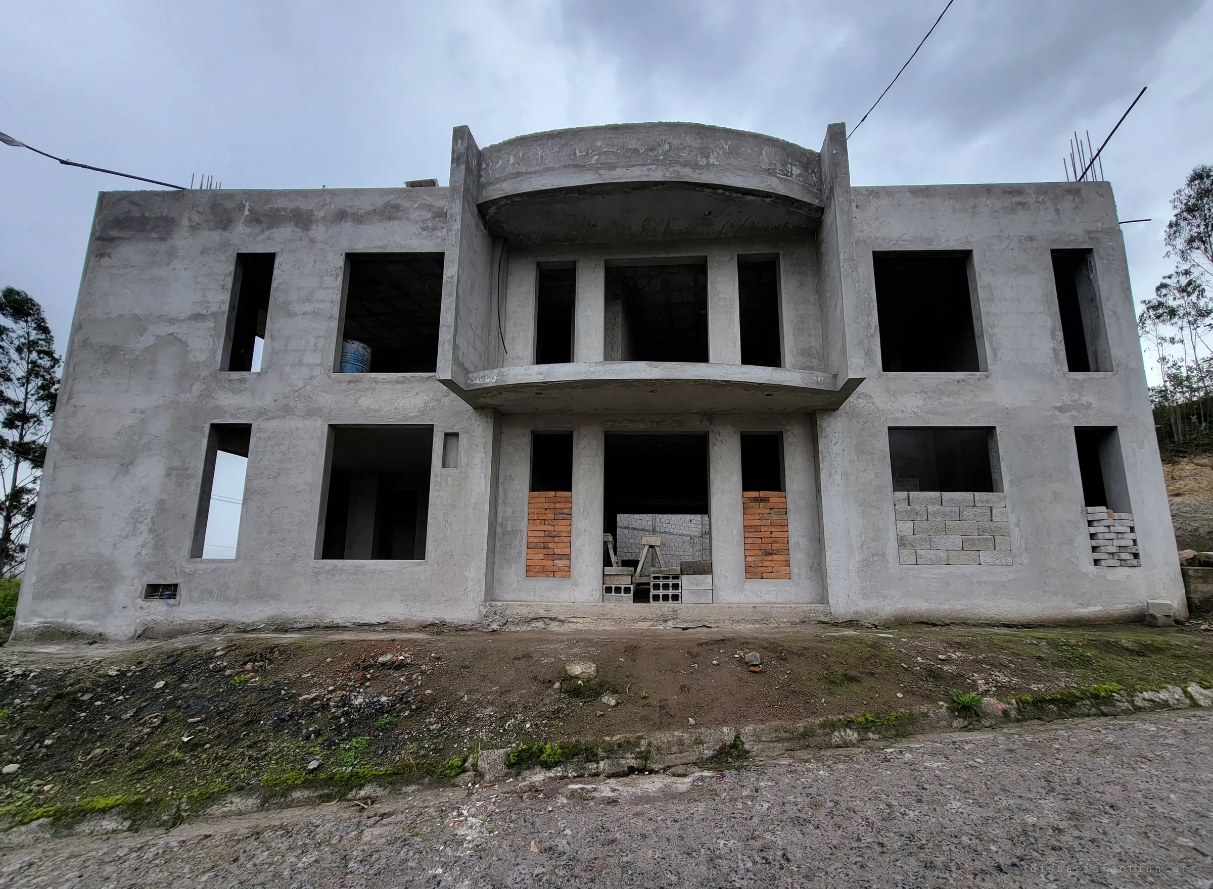 The bare concrete front of an unfinished two-storey house under a grey sky, its curved balcony empty and window openings half bricked up. Sawhorses and cinder blocks still sit in the doorway as if the builders just stepped out.