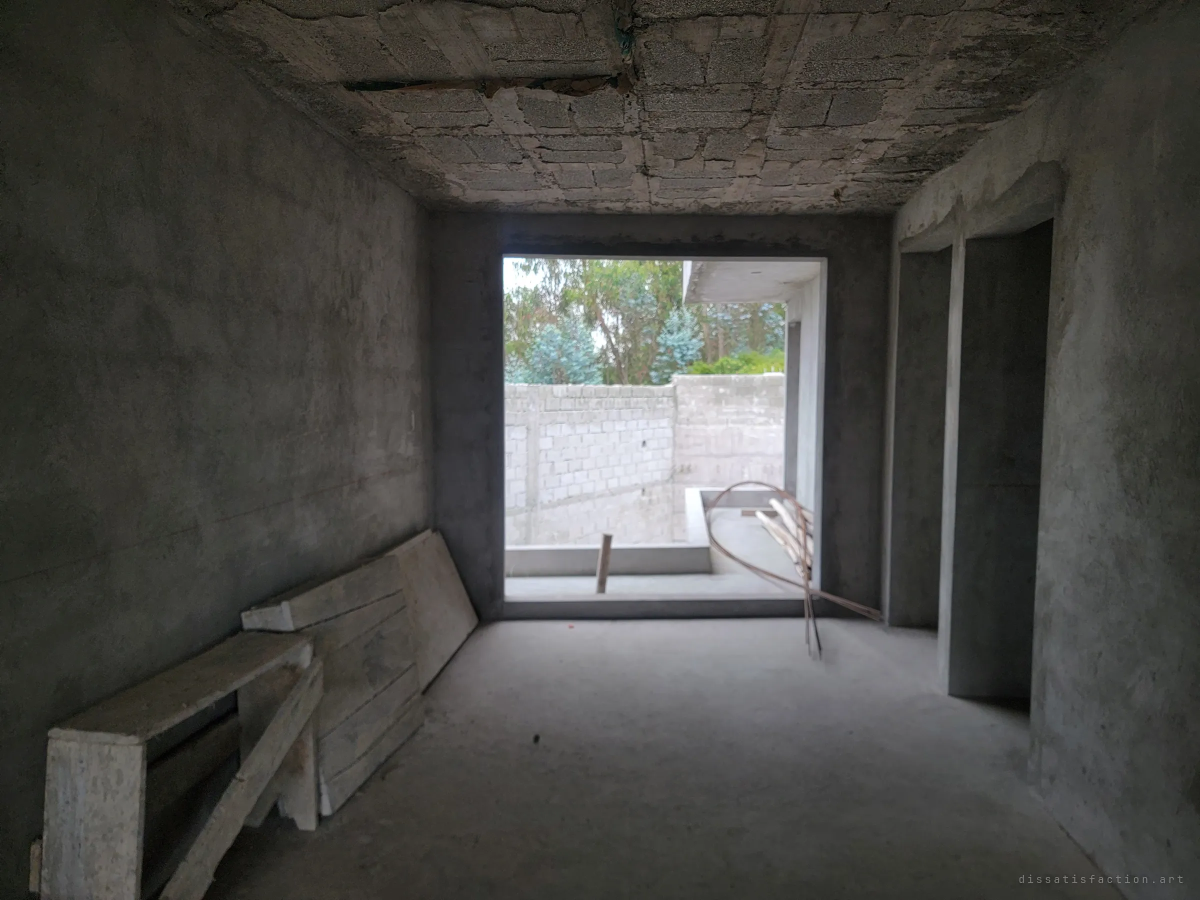 A dim concrete hallway ends in a wide opening onto a walled courtyard and trees. Old formwork planks lean against the wall and a coil of copper pipe sags in the doorway.