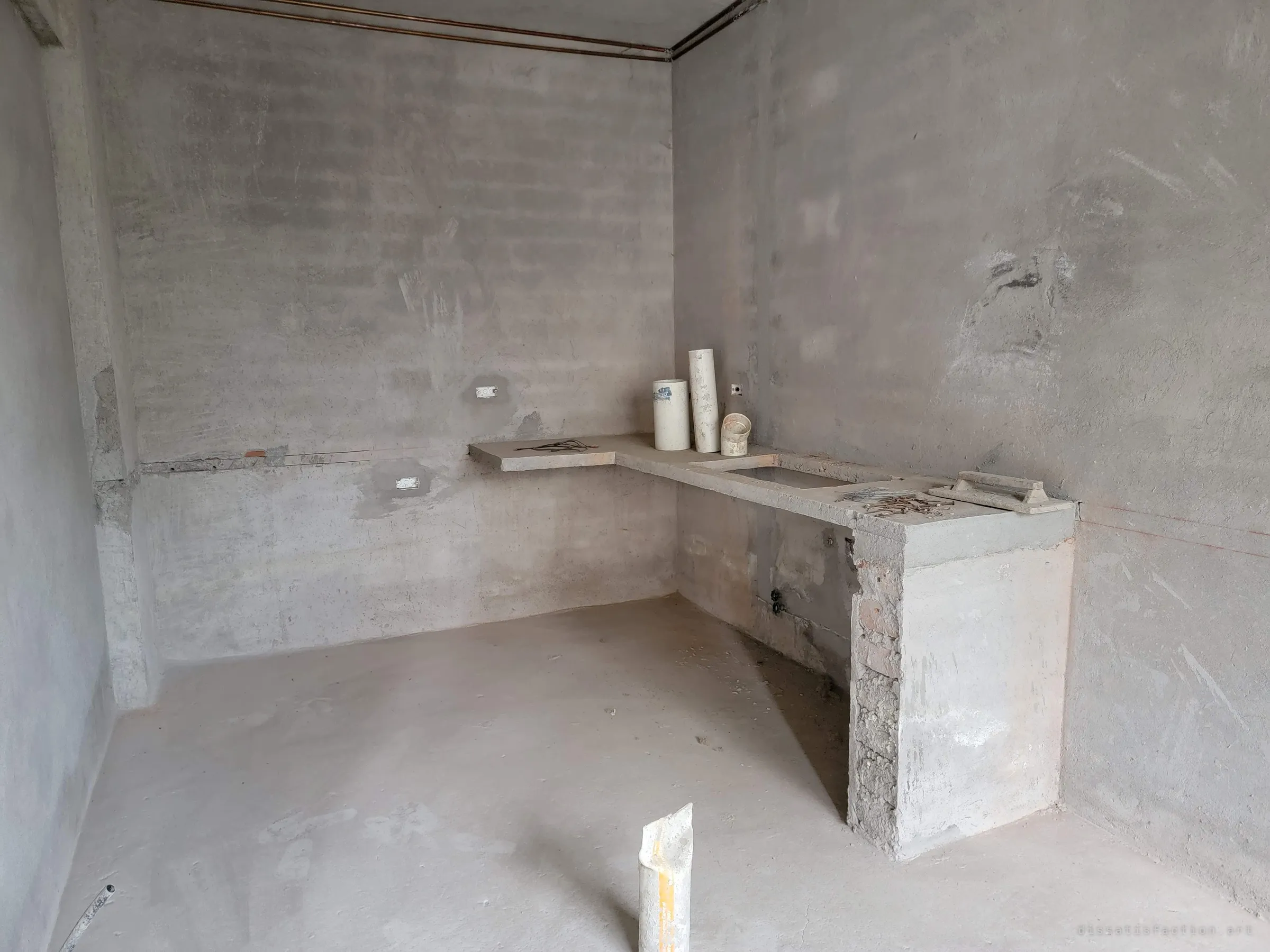 A bare room with a cast concrete counter and sink cut-out where a kitchen was meant to go. PVC pipe offcuts and a trowel are still on the counter.