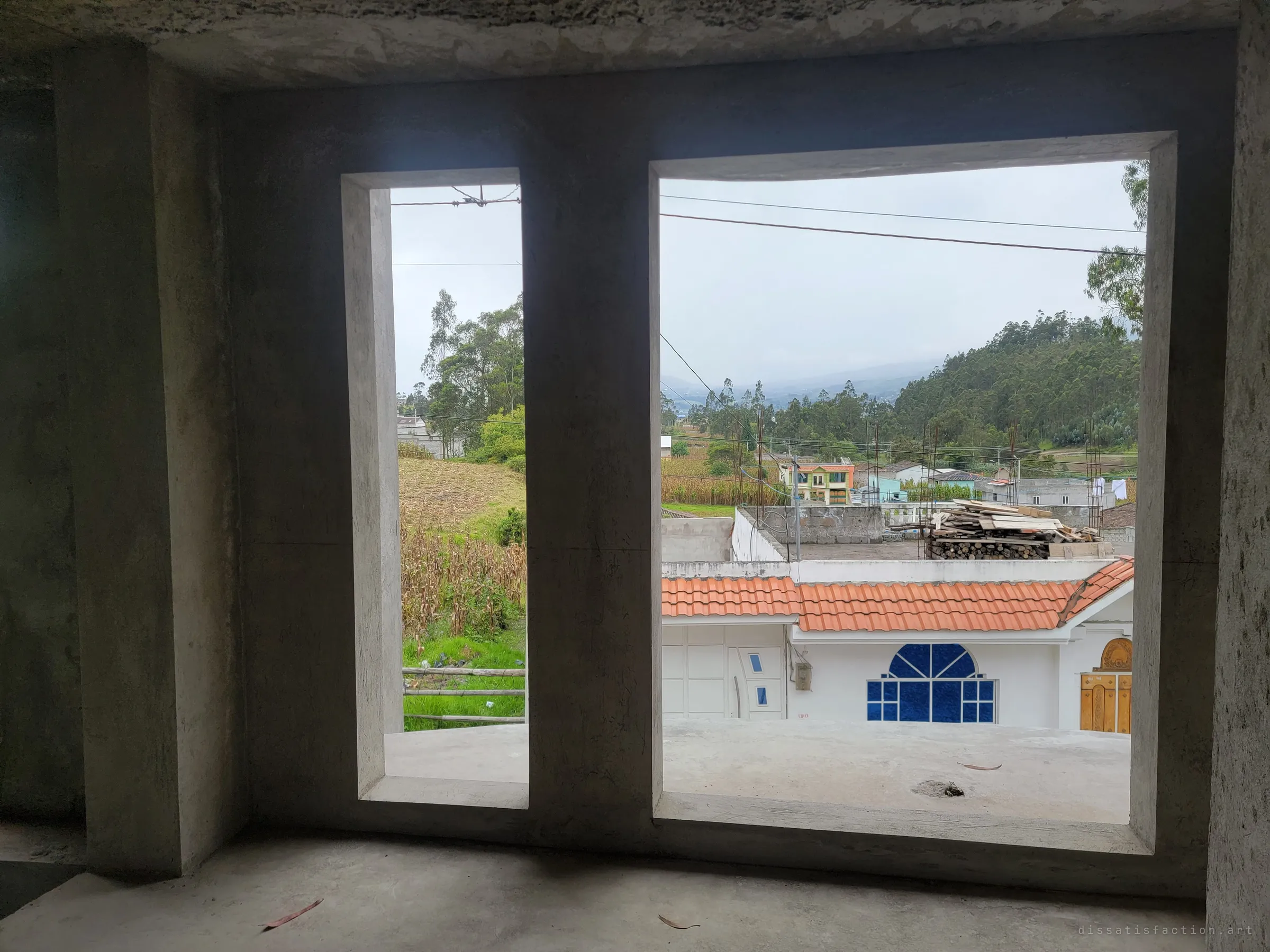 Two tall empty window openings frame the view from the upper floor: a neighbour's tiled roof, cornfields and misty eucalyptus hills. The raw concrete frame makes the finished houses outside look like another world.