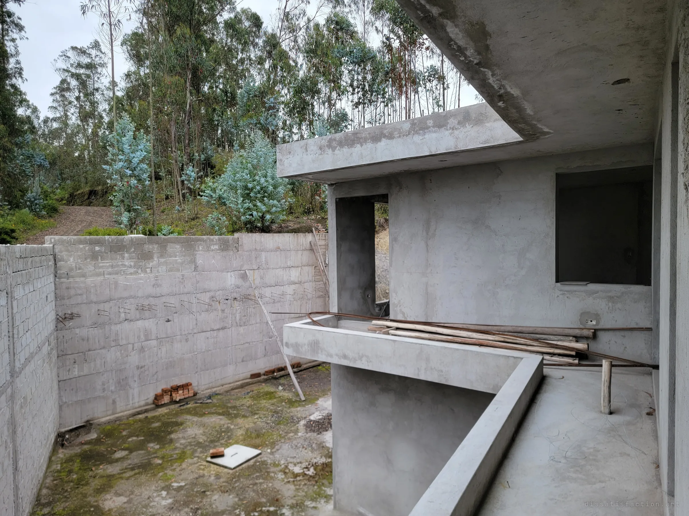 A cantilevered concrete slab juts over a mossy sunken courtyard, walled in by cinder block and pale eucalyptus. Poles and rebar lie across the half-built balcony.
