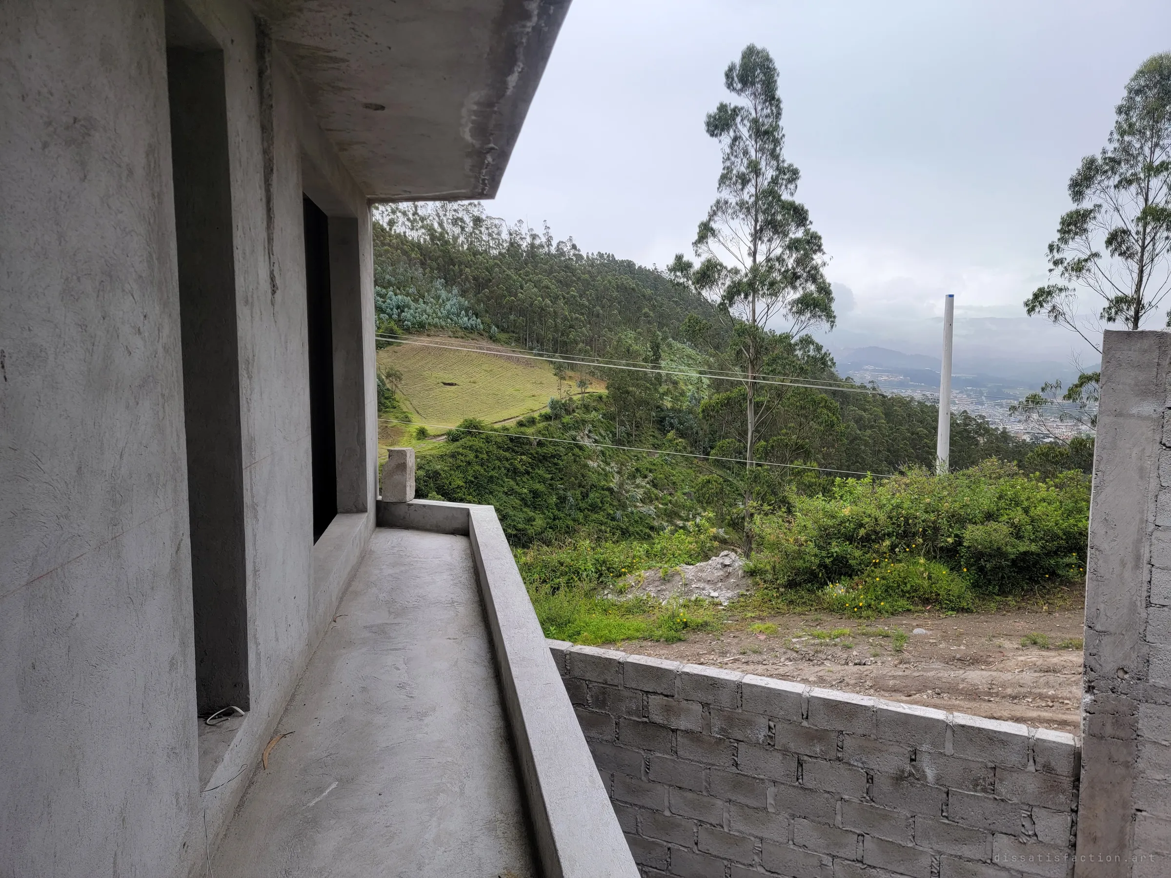 A bare concrete balcony runs along the unfinished upper floor, looking out over eucalyptus slopes and a misty valley. The raw grey shell framed against open hillside sets the half-built mood of the house.