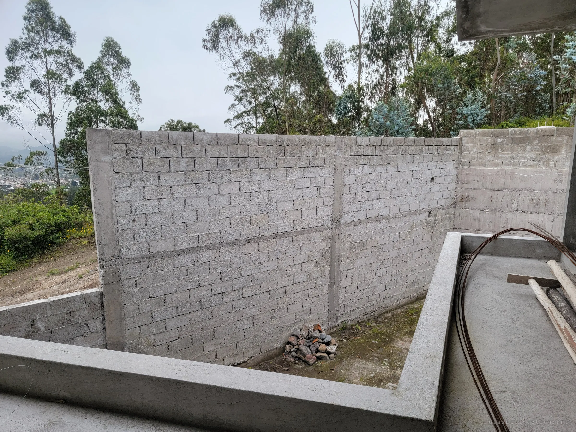 A tall cinder-block retaining wall encloses an empty yard beside the house, with a small pile of stones at its foot and coiled rebar on the slab. Eucalyptus trees crowd in above the unfinished boundary.