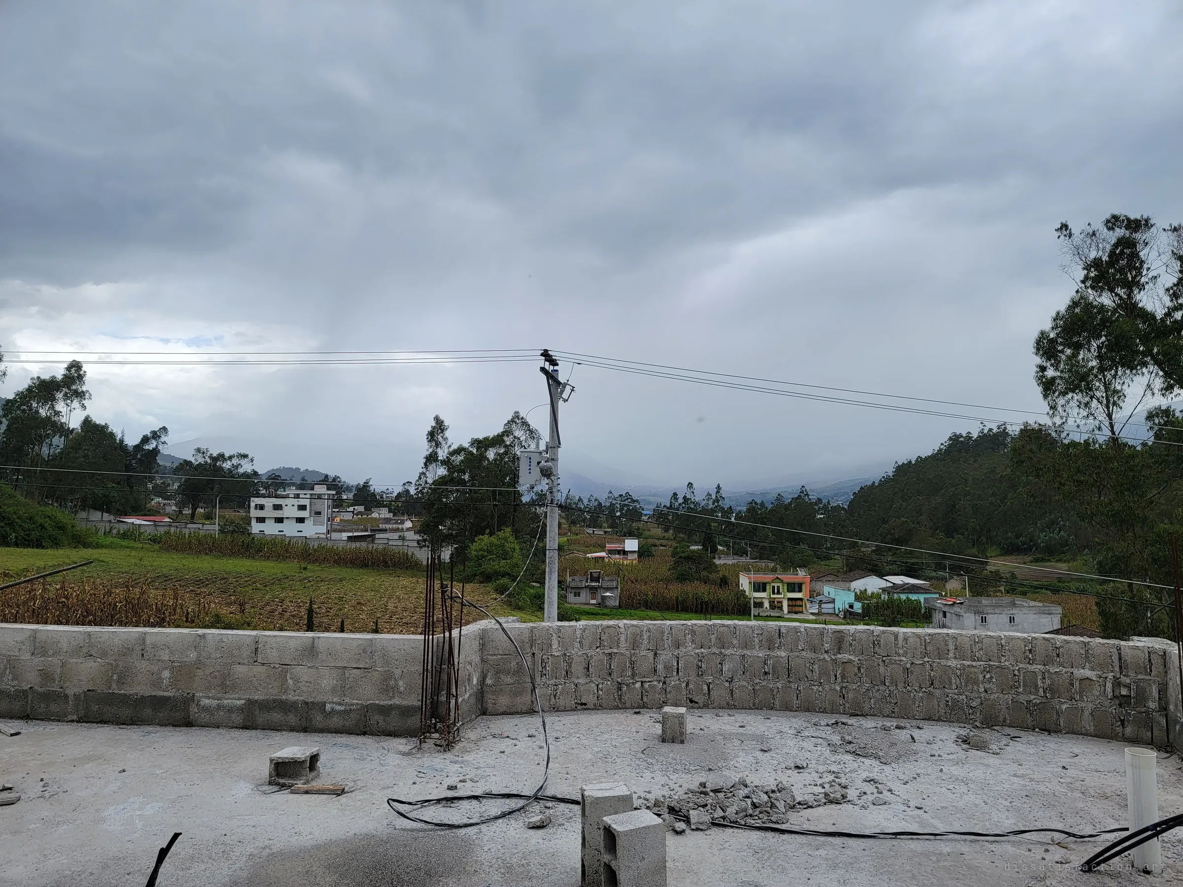From the open roof slab, a low block parapet looks over cornfields and scattered village houses under a heavy grey sky. Loose blocks, rubble and cables lie across the unfinished roof.