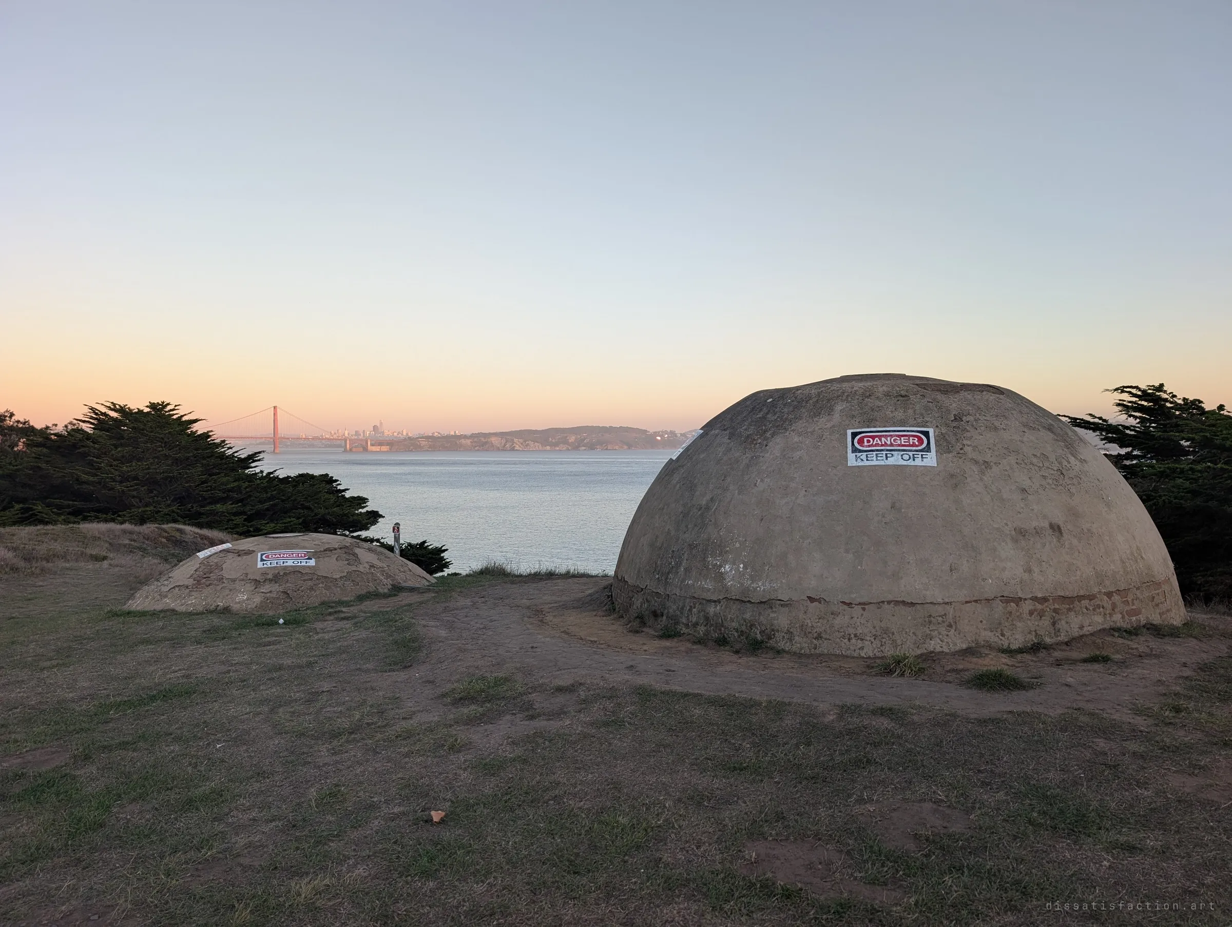Two weathered concrete domes marked with danger warning signs sit on a grassy bluff at dusk, water and a distant bridge behind them. The domes are the capped tops of old coastal defence works.