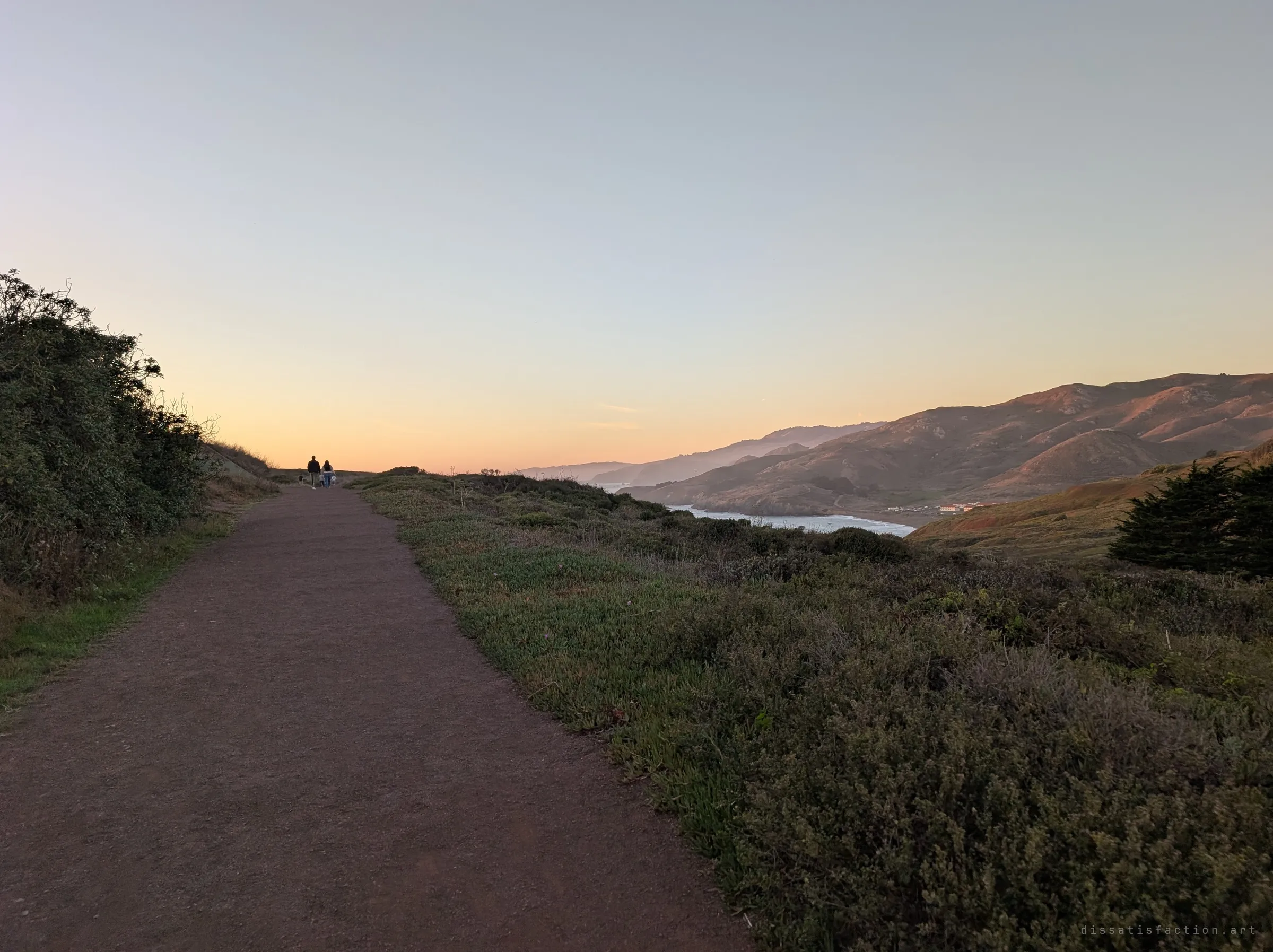 A dirt trail runs along a scrubby ridge toward two distant walkers at sunset, hills and a cove falling away to the right. It sets the approach to the site.