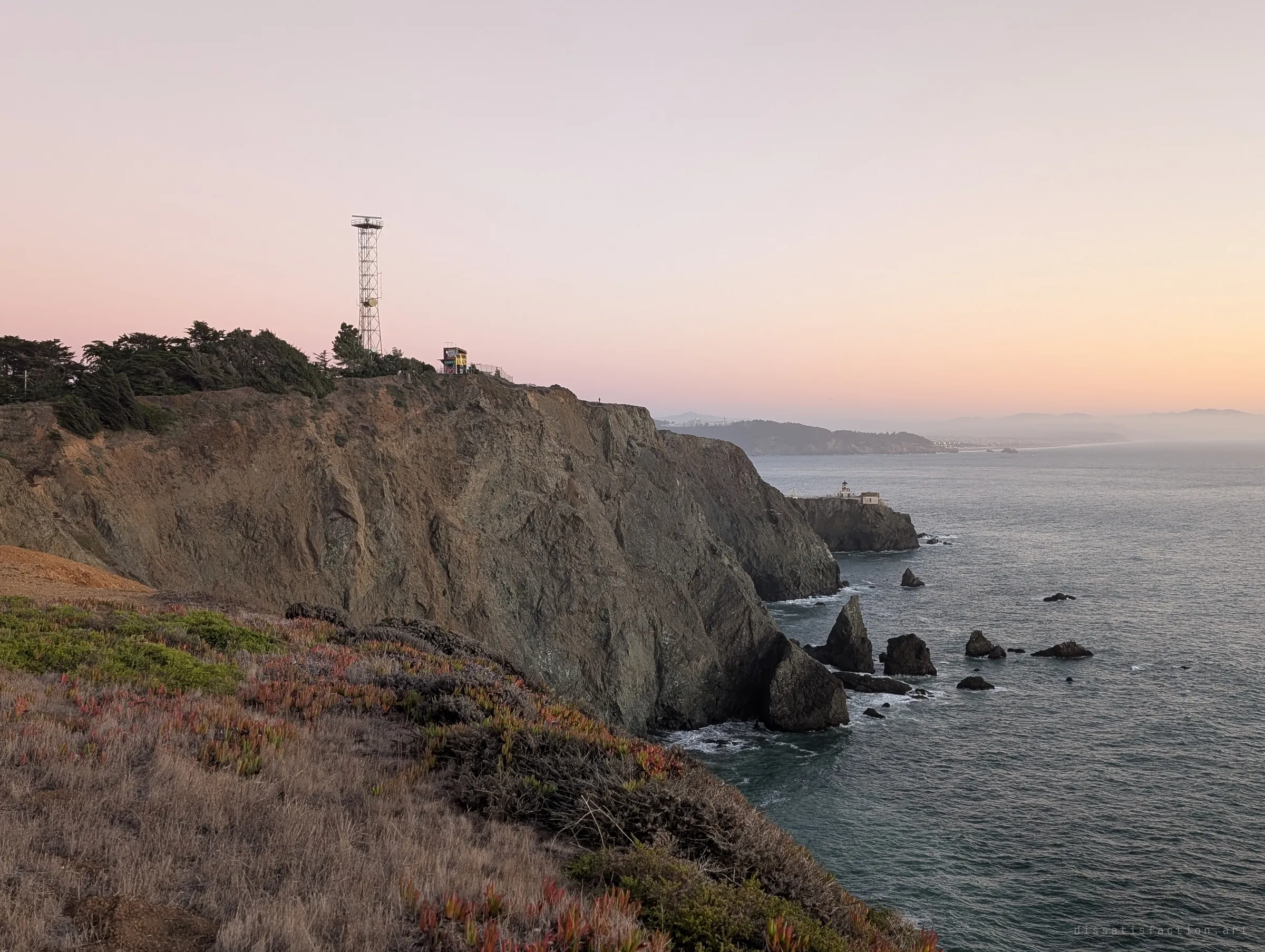 Sheer cliffs drop into the sea under a pink sky, a lattice tower and small lookout on the crest and a tiny lighthouse on the point below. It shows how exposed the headland is.