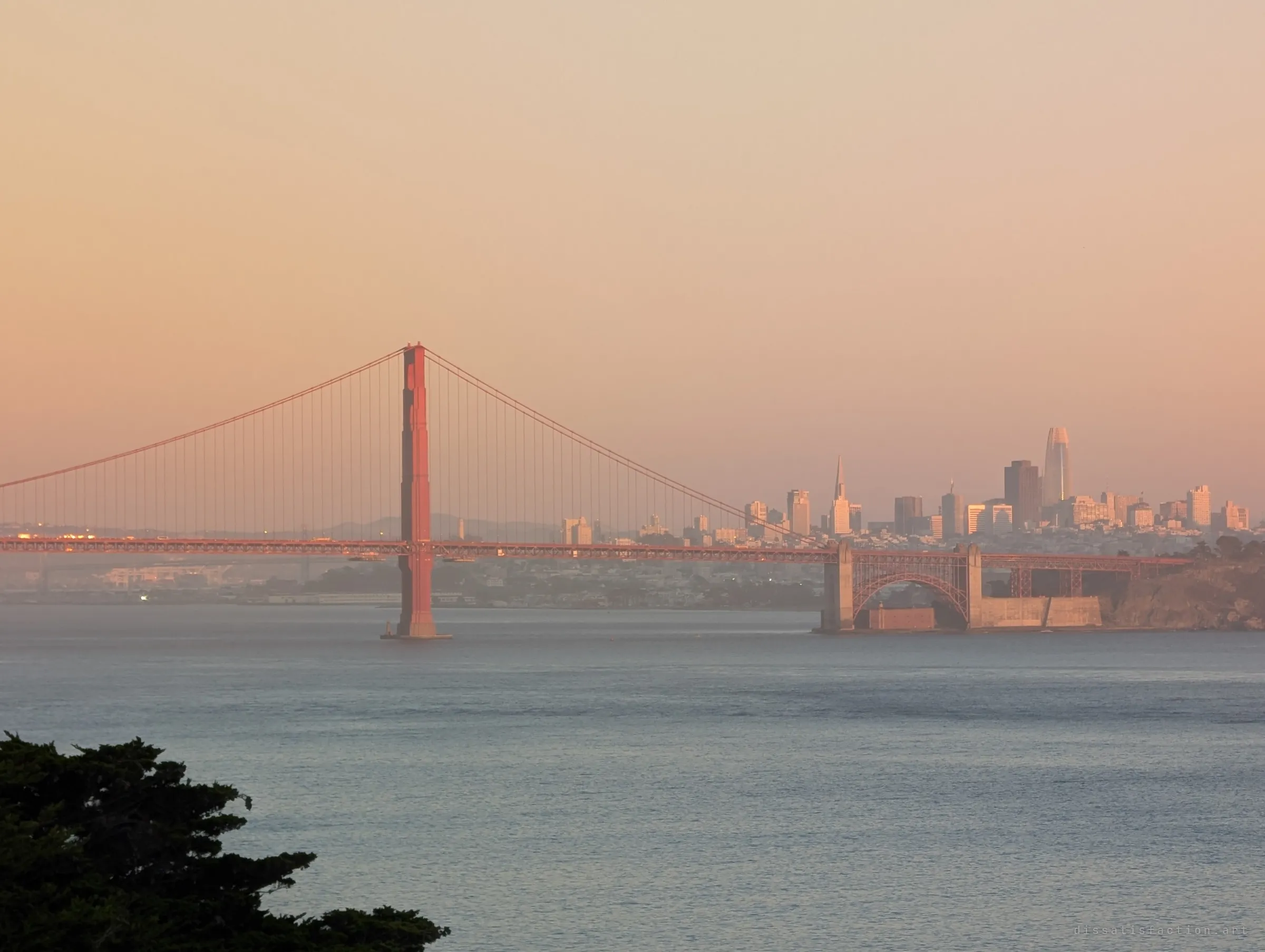 A red suspension bridge tower and a hazy city skyline glow in late orange light over calm water. A view from the high ground above the site.