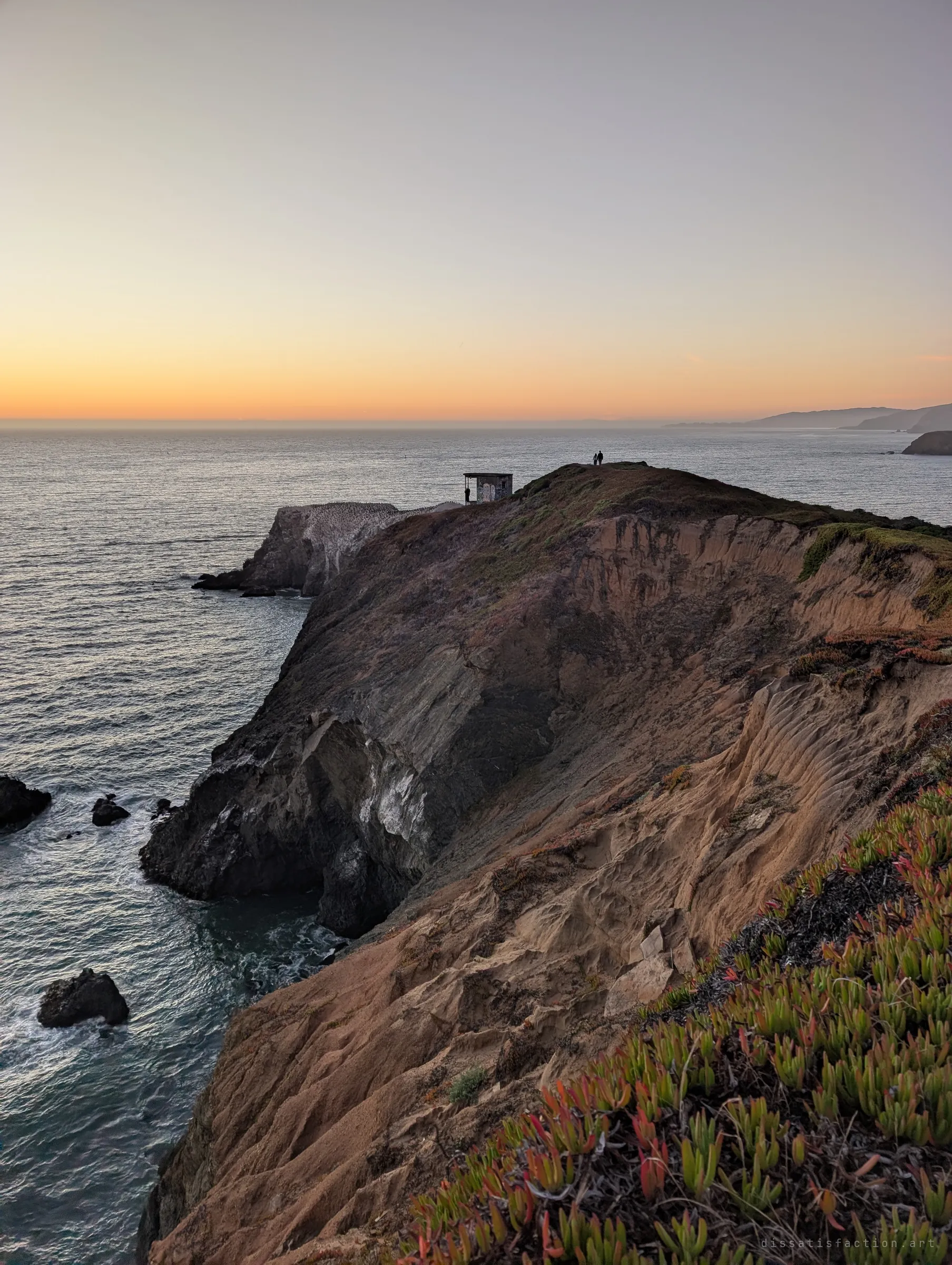 A crumbling sandstone ridge leads out to a small ruined lookout on the edge of the cliff, with two figures silhouetted against the sunset sea. The lookout is one of the site's scattered outposts.