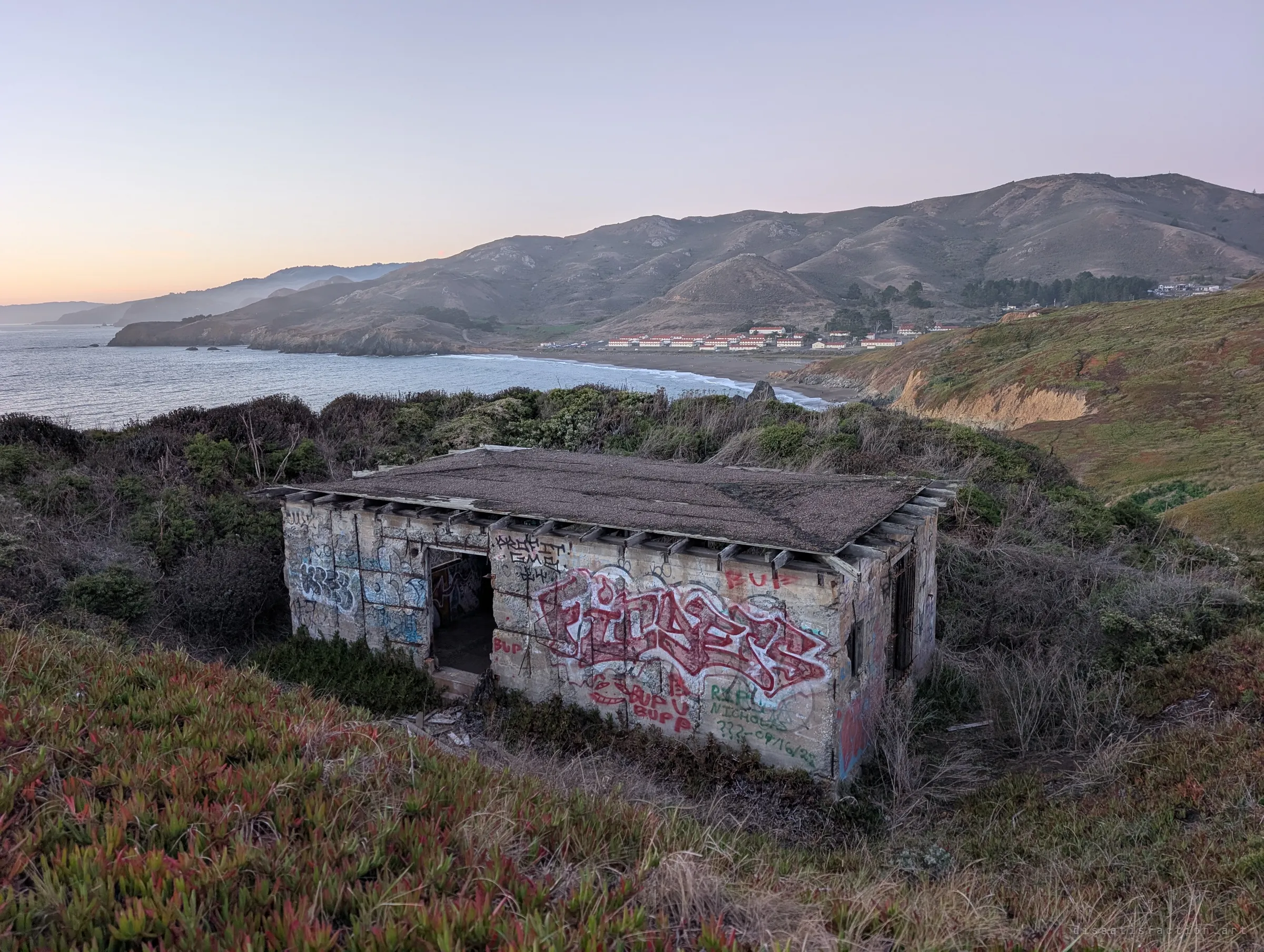 A small concrete hut covered in graffiti sits in scrub above a curving beach, its tar roof sagging over broken rafters. It is one of the abandoned support buildings scattered across the hillside.
