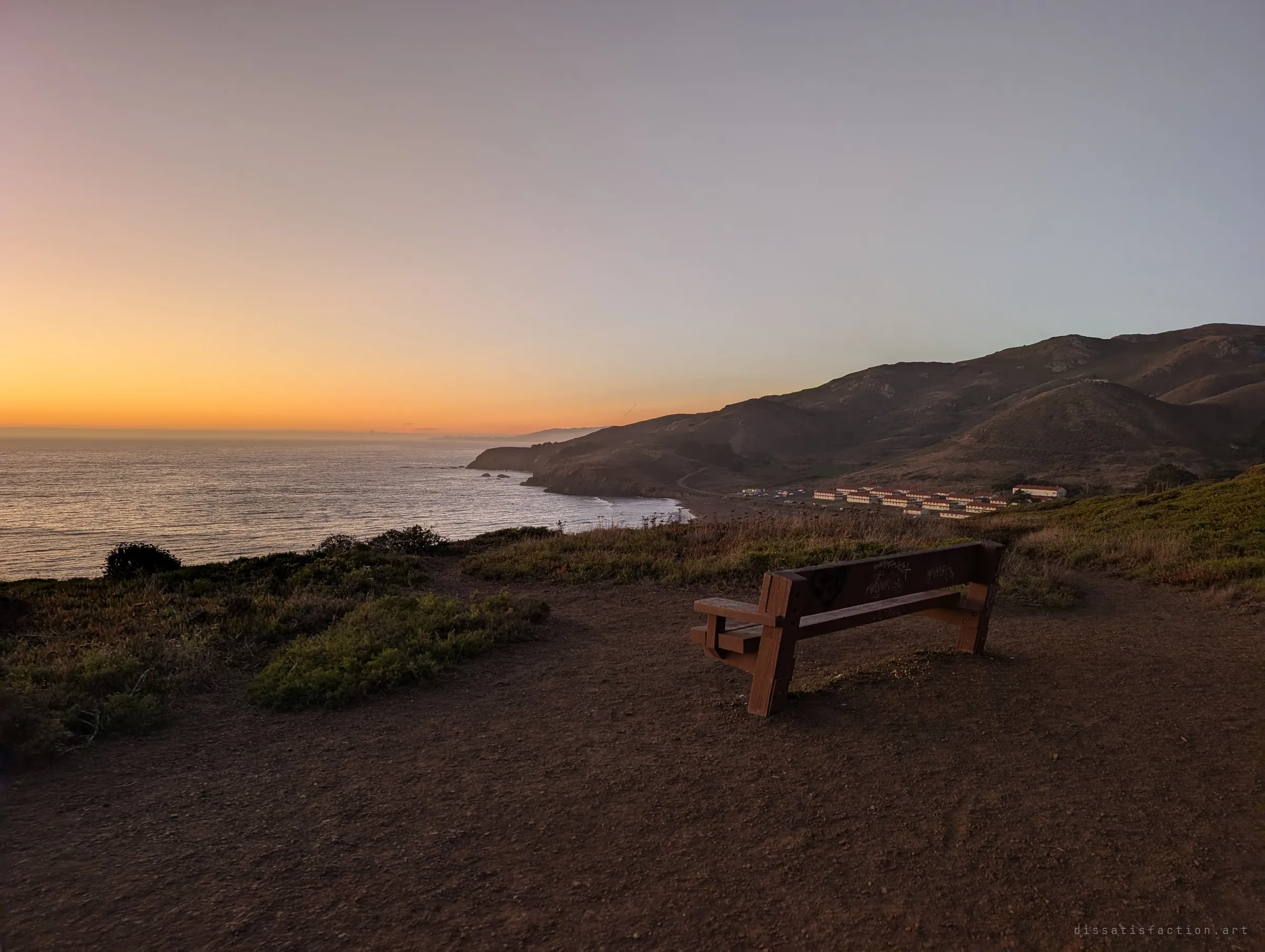 A tagged wooden bench faces the ocean at sunset from a dirt clearing, hills and a beach curving away to the right. The quiet overlook at the edge of the site.