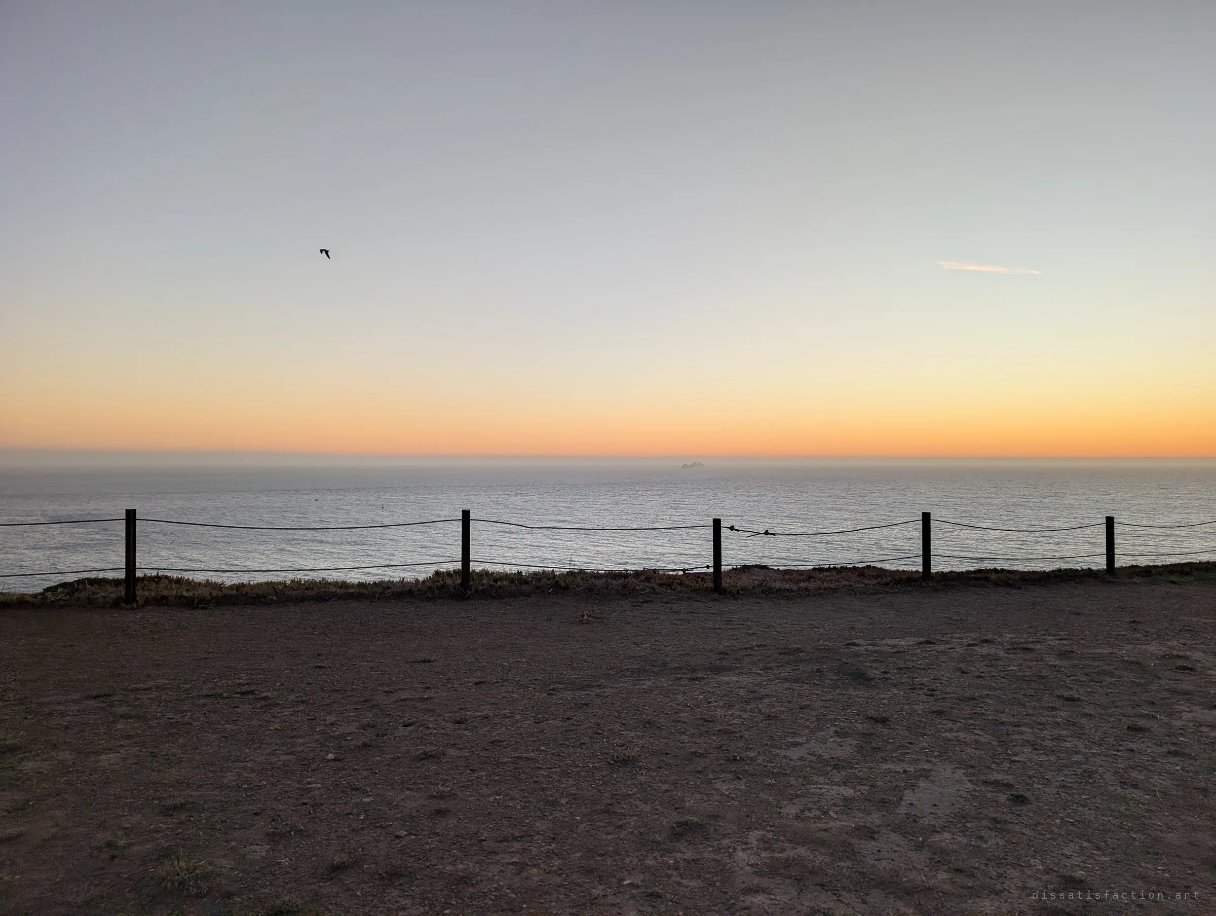 A simple rope fence runs along a clifftop above open ocean, a lone bird crossing the dusk sky. The empty edge beyond the fortifications.