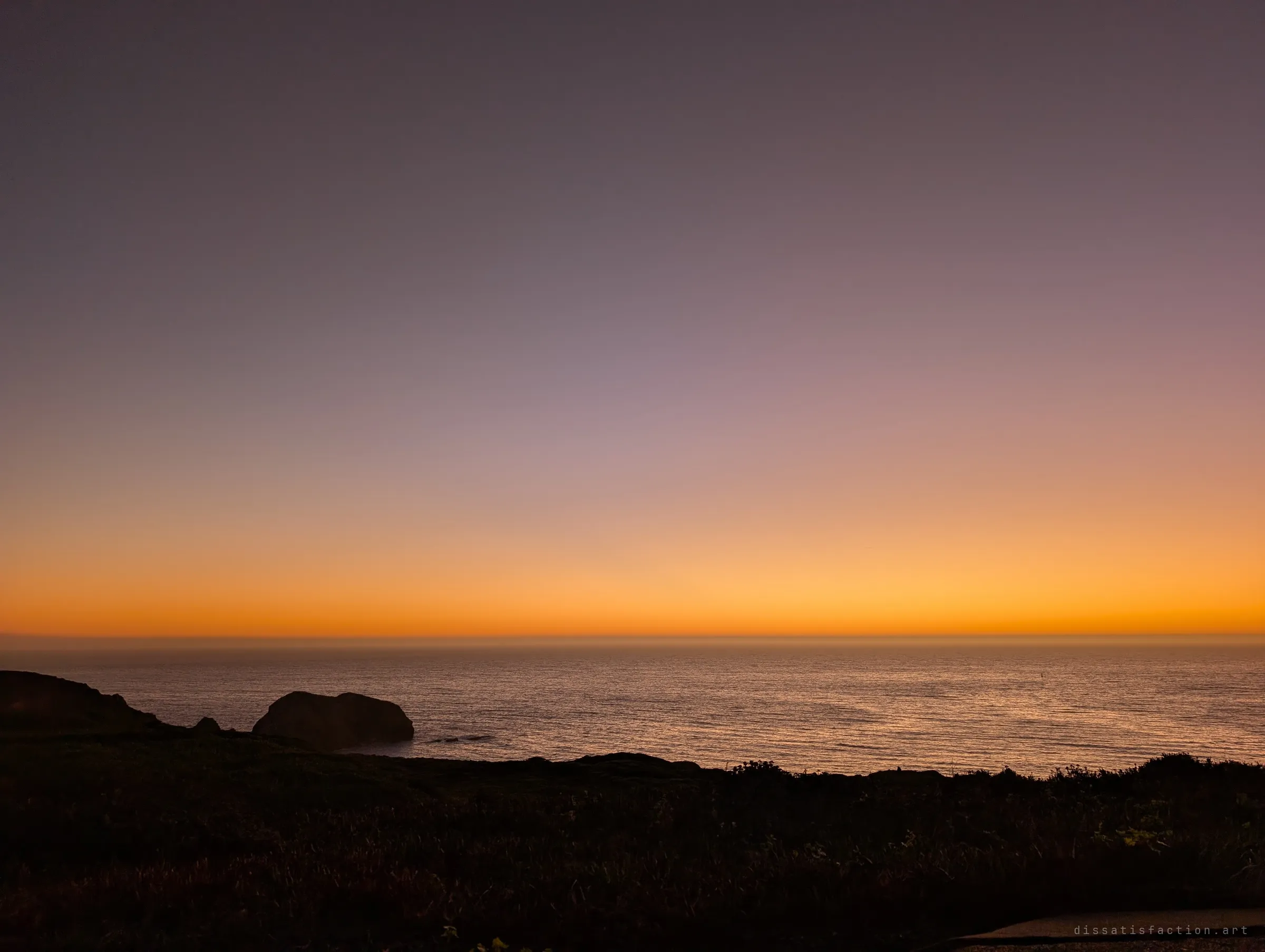The sea glows orange under a violet afterglow, a dark rock and the clifftop silhouetted in the foreground. The last light over the headland.