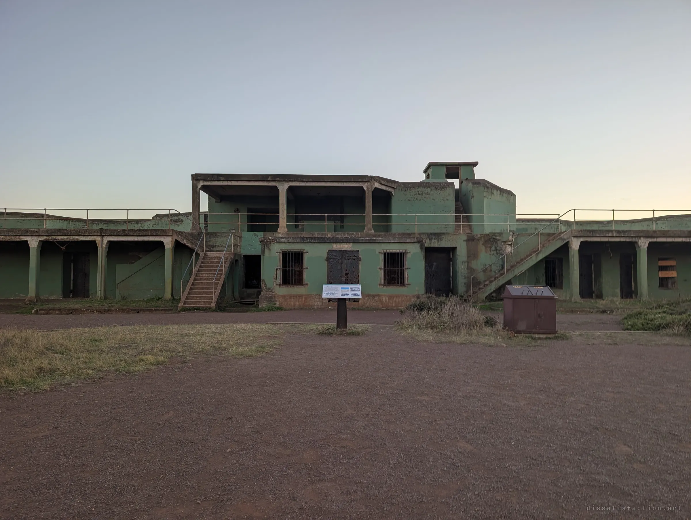 A two-storey green concrete gun battery with barred windows, steel doors and external stairs stands across a gravel yard. The main structure of the site, seen head-on.