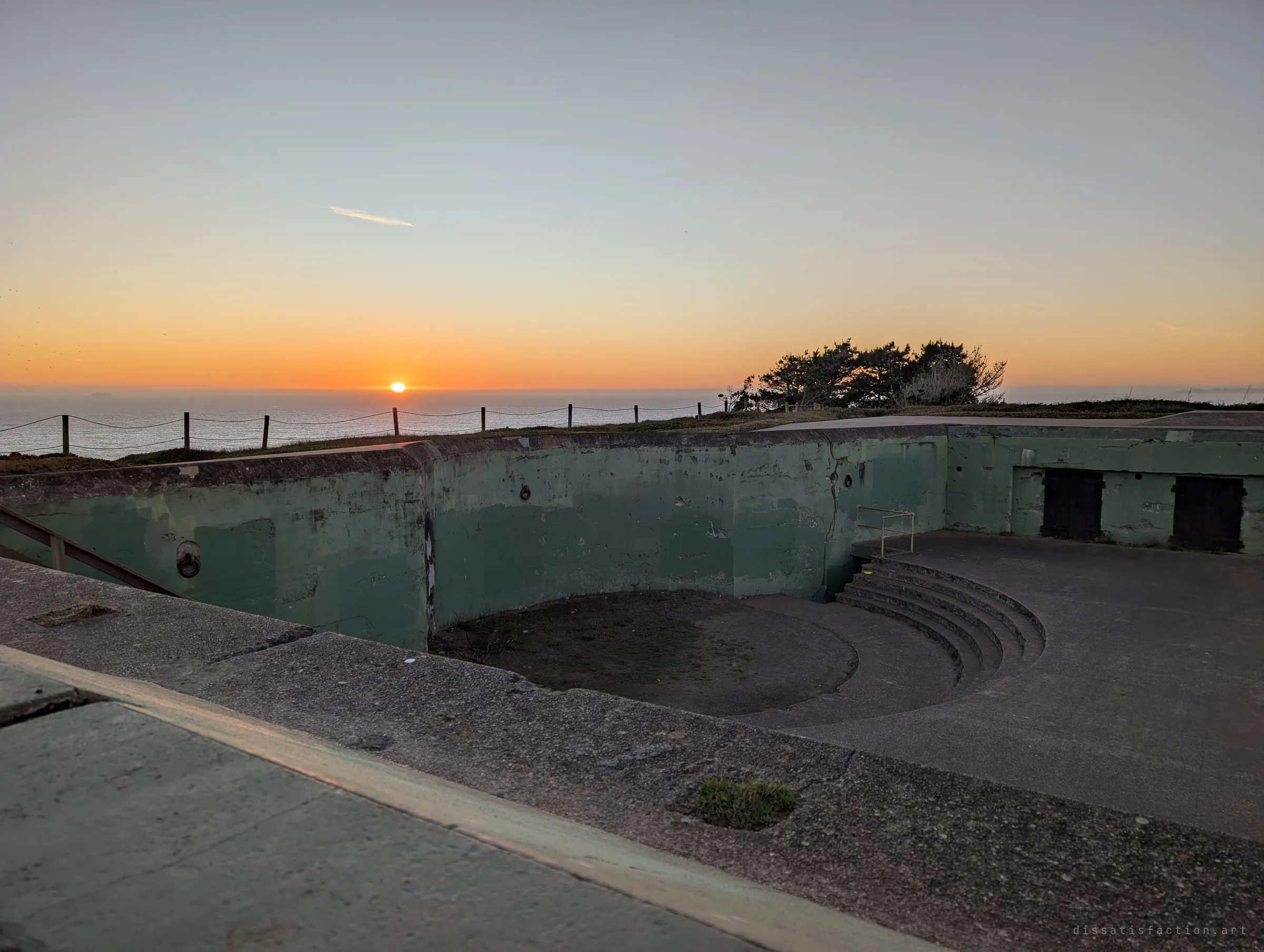 A circular gun pit with curved concrete steps and green walls opens toward the setting sun over the sea. The empty mount where a heavy gun once turned.
