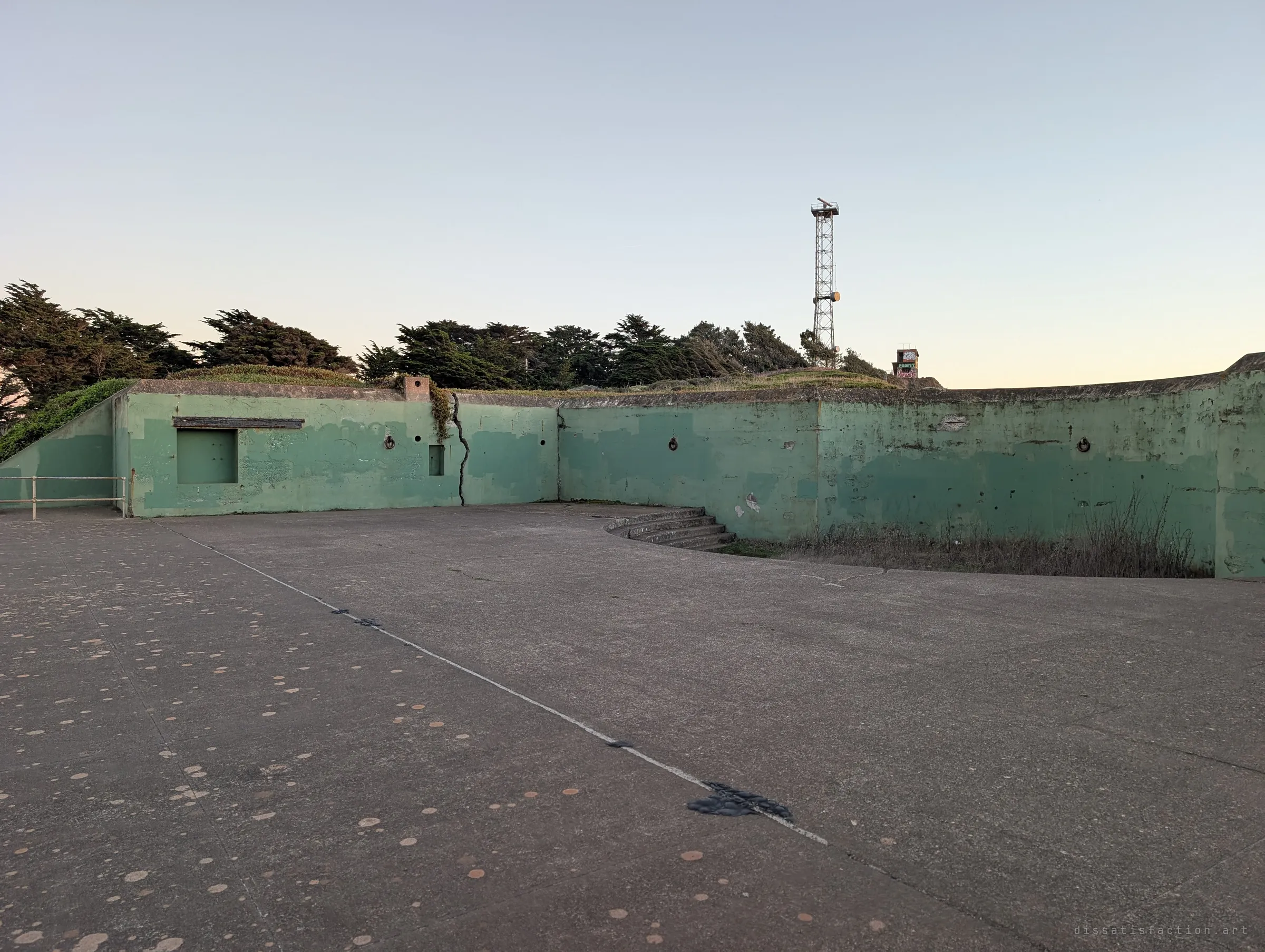 A wide concrete gun platform with cracked green walls and mooring rings, a lattice tower rising behind the trees. It shows the scale of the emplacement floor.
