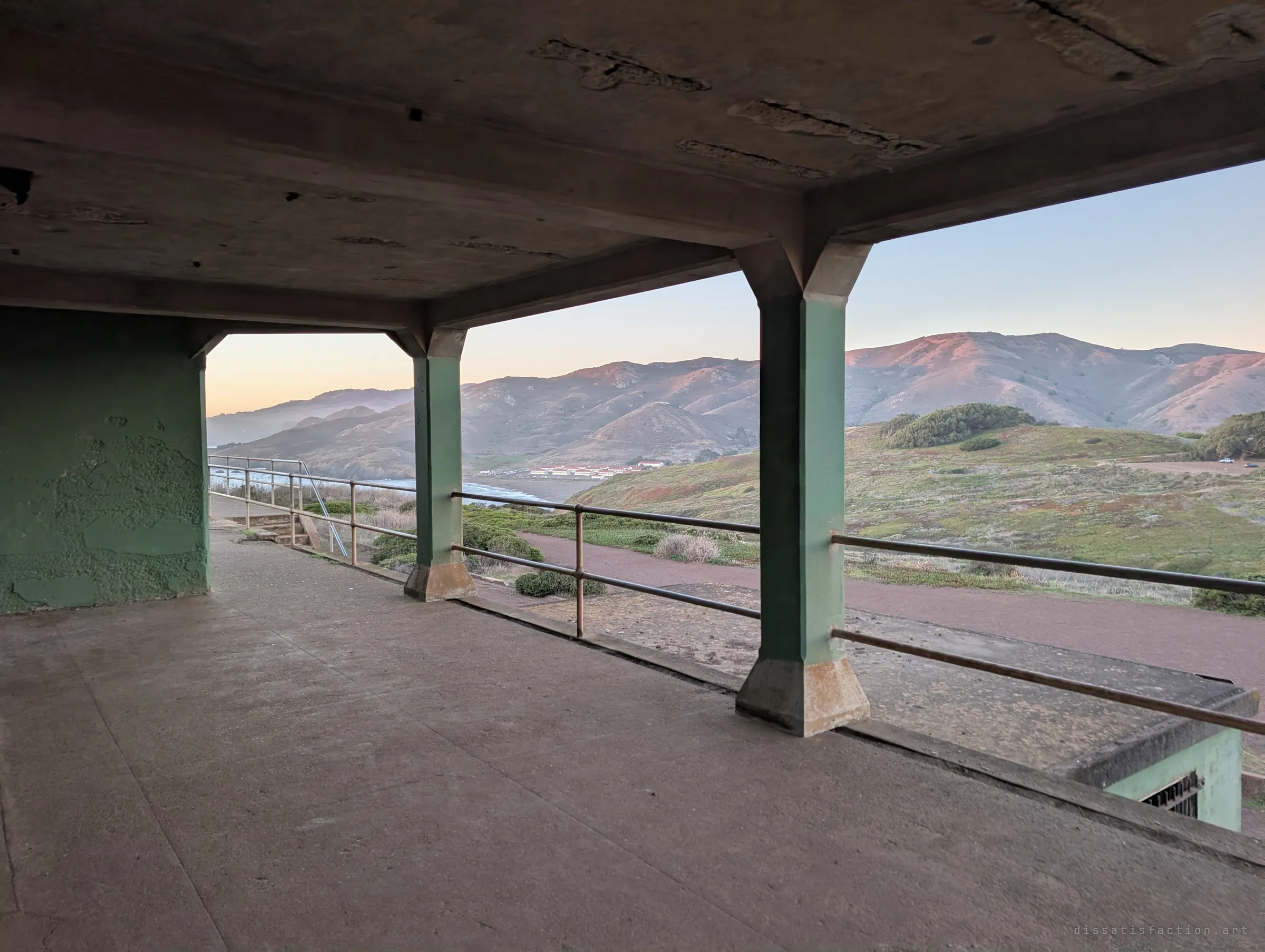A green-painted concrete gallery with square pillars frames a sunrise view over grassy hills and a far-off shoreline. The open, railed deck shows how the structure was built to watch over the landscape.