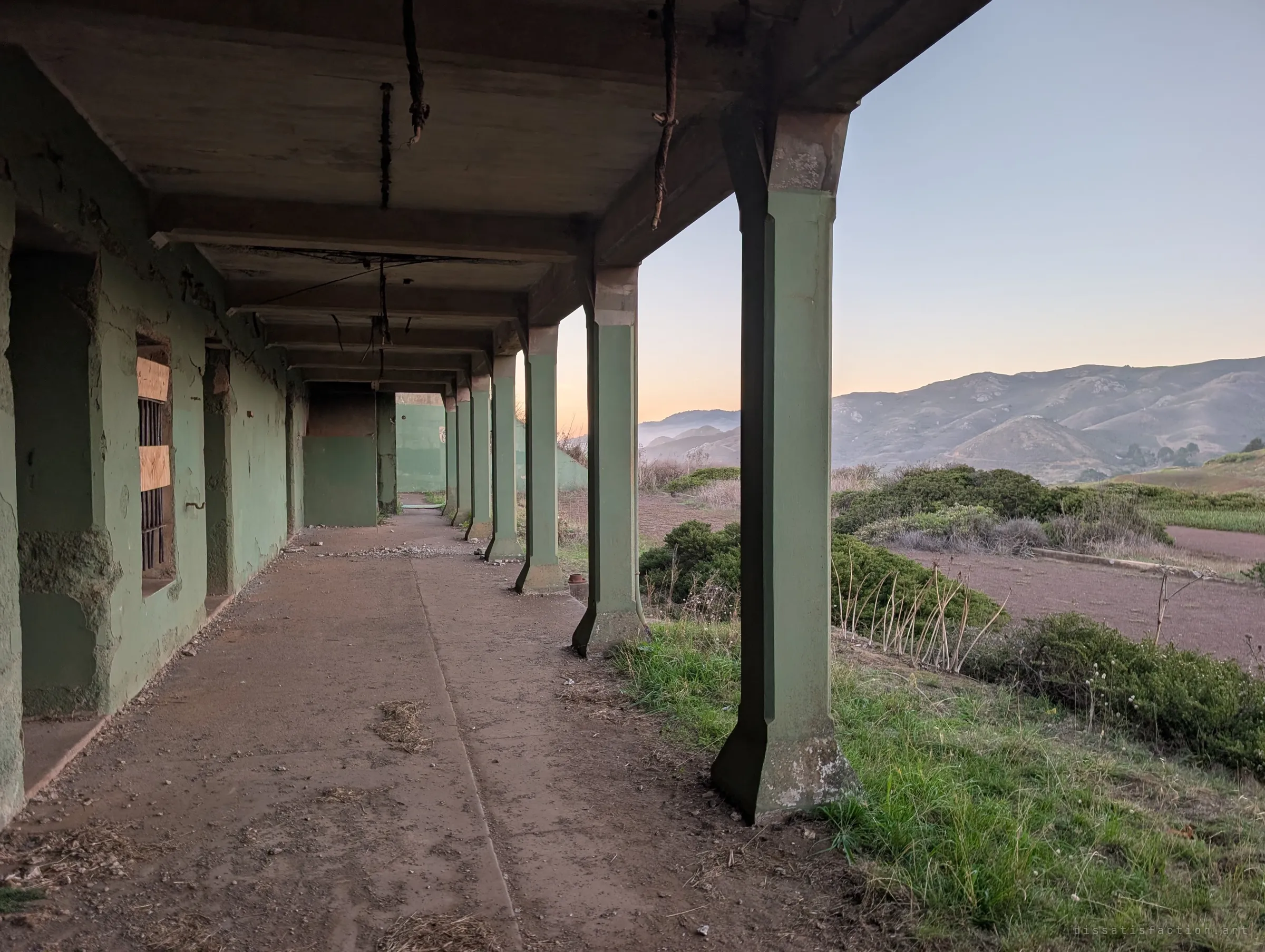 A long covered walkway of green concrete pillars runs past barred, boarded windows, with dry scrub and hills glowing at dawn beyond. Rusted fittings hang from the ceiling where lights once were.