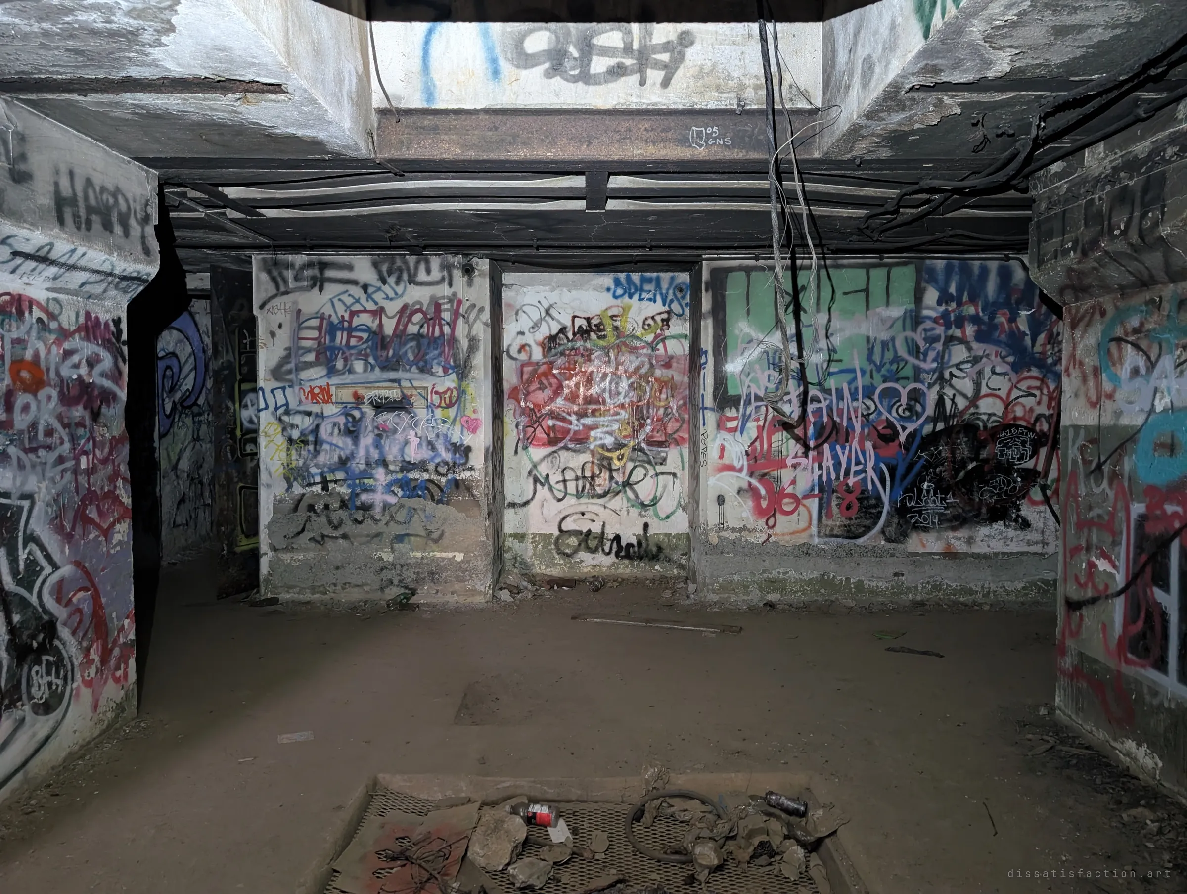 An underground concrete room covered wall to wall in layered graffiti, with loose cables drooping from a light shaft and a rubble-strewn grate in the floor. Decades of visitors have left their marks on every surface.
