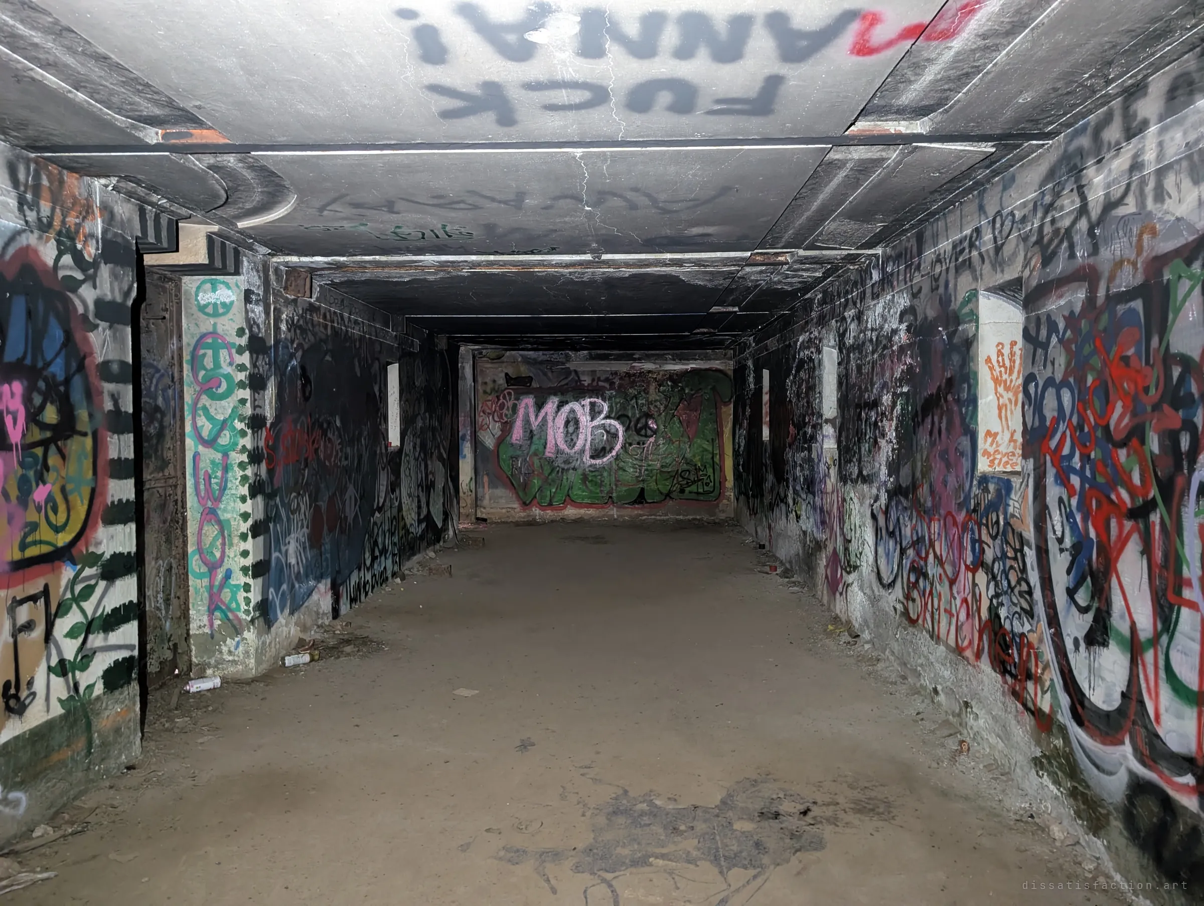 A long, low concrete corridor with graffiti on the walls and ceiling leads to a far wall with a large pink-and-green piece. Discarded spray cans lie along the dusty floor.