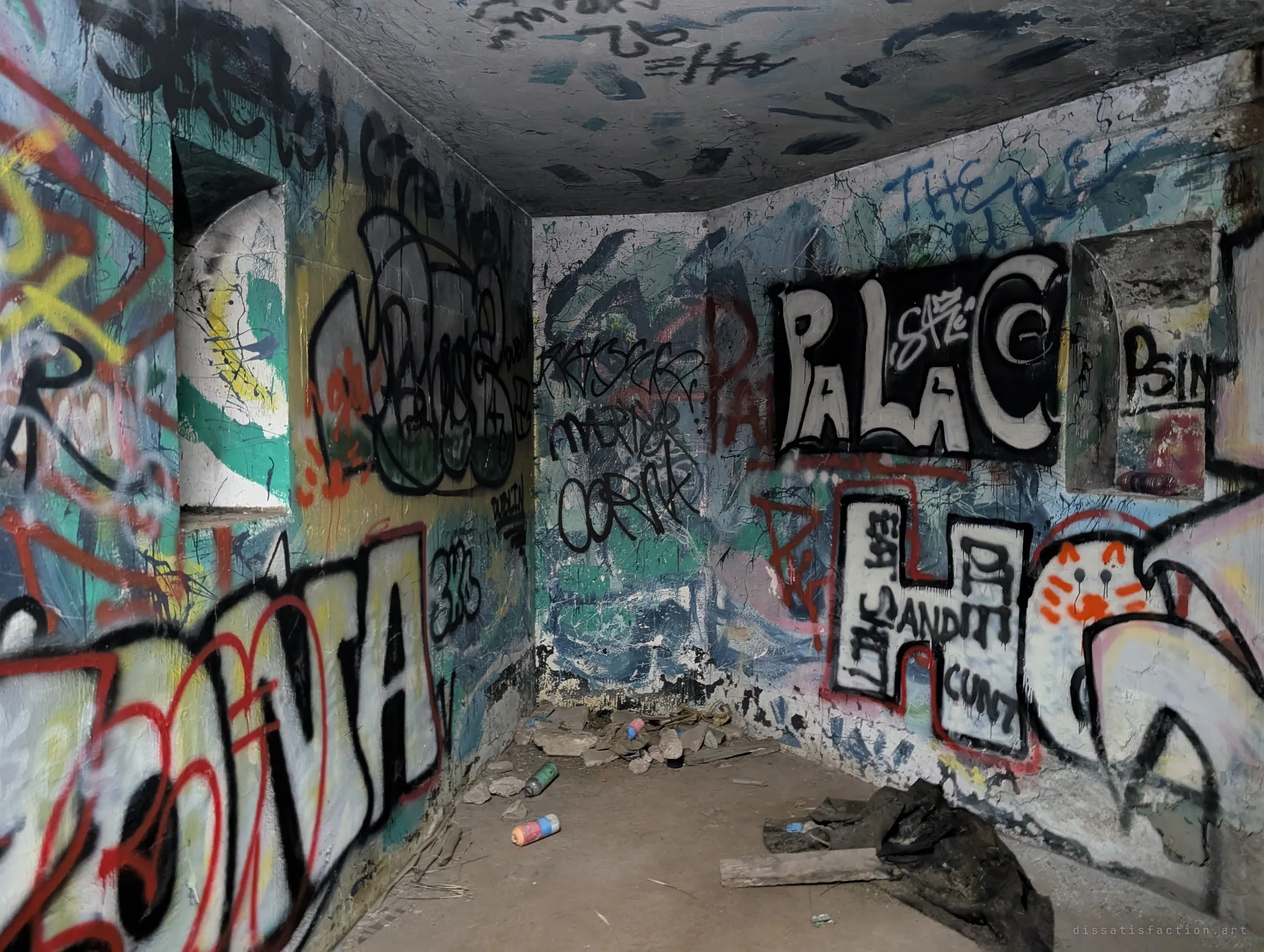 A small concrete chamber with arched wall niches, every inch painted with bold graffiti letters and a cartoon cat. Empty spray cans, rubble and a rotting tarp sit on the floor.