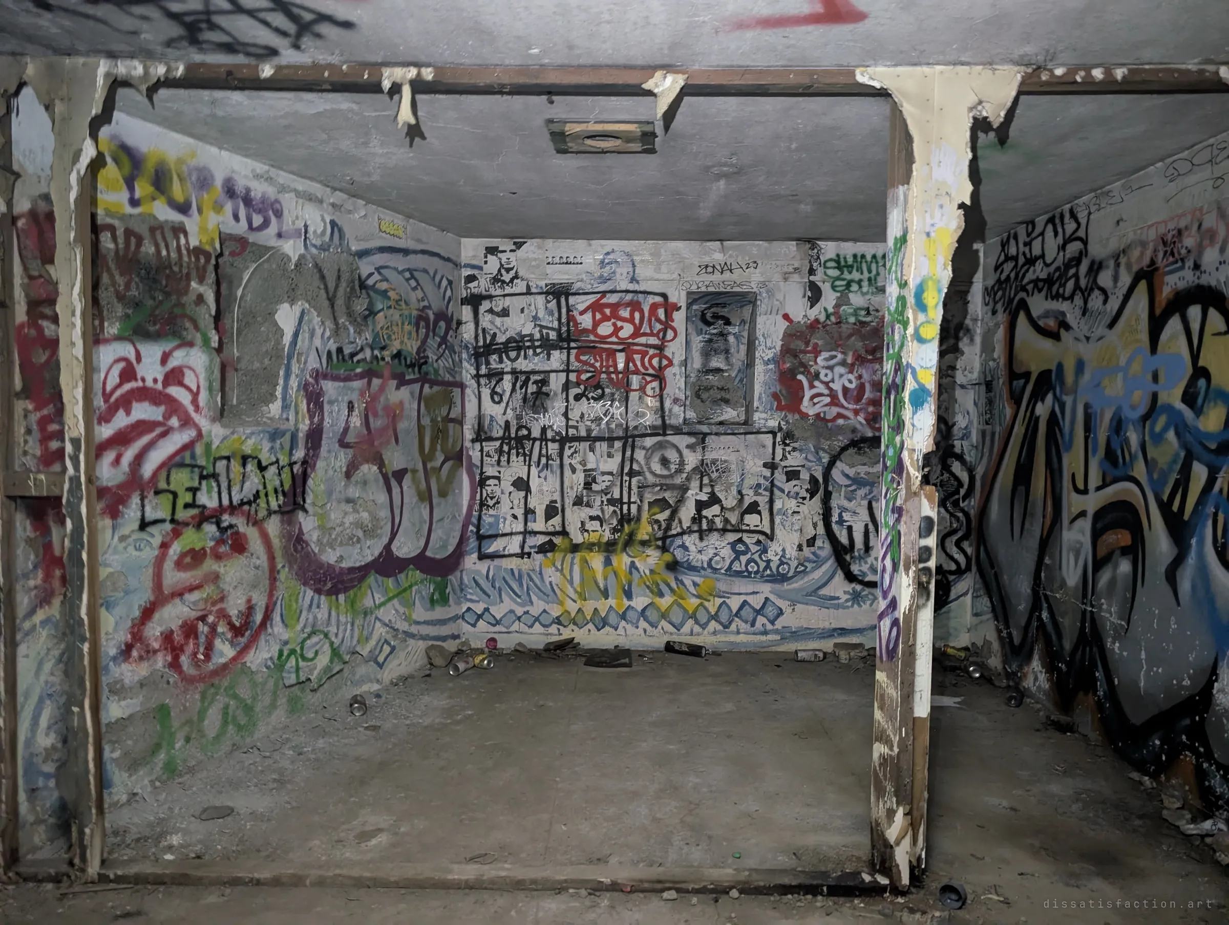 A concrete room framed by peeling wooden posts, its back wall covered in pasted-up portrait posters, tags and a painted wave pattern. Empty cans are scattered across the bare floor.