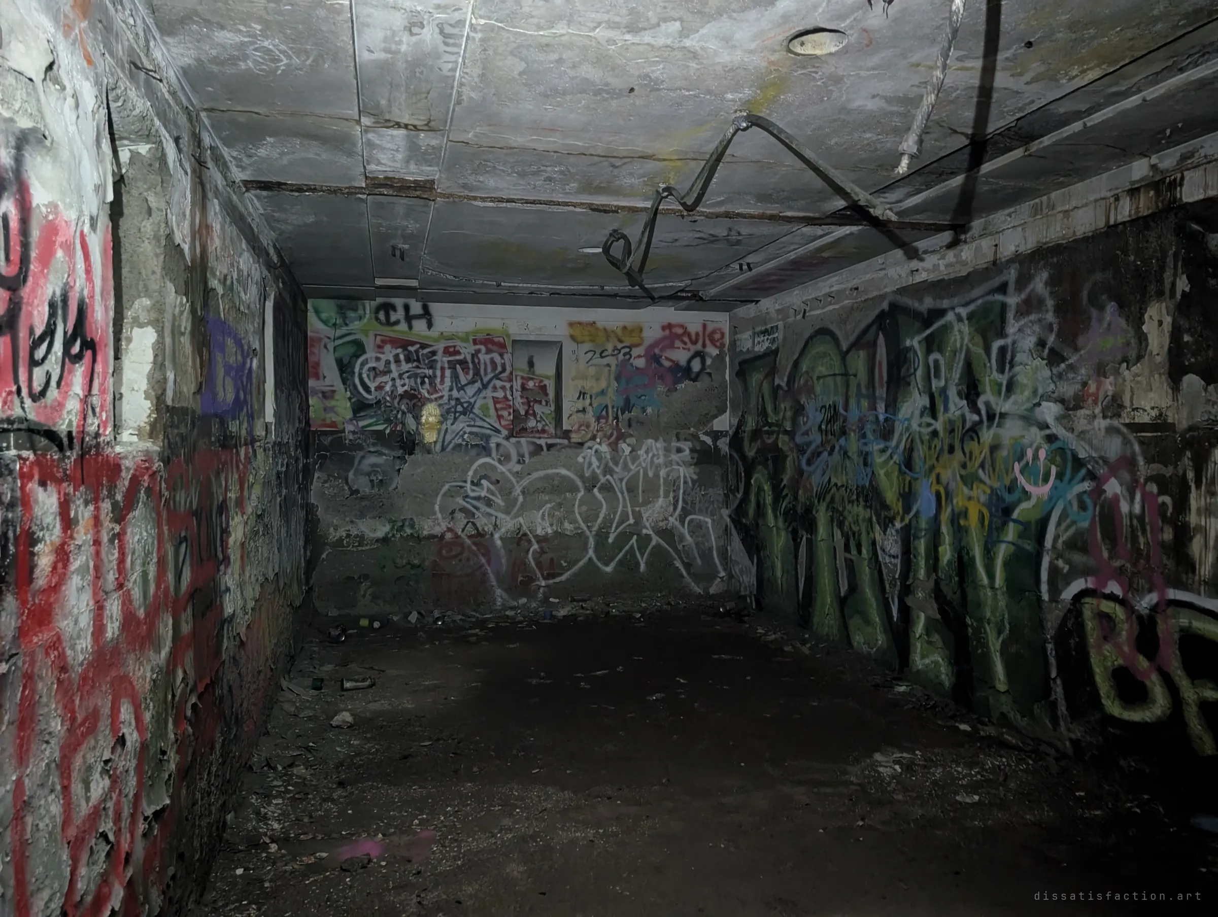 A dim graffiti-covered chamber lit by a flashlight, with a twisted cable hanging from the cracked ceiling slab. Peeling paint and layered tags show years of neglect.